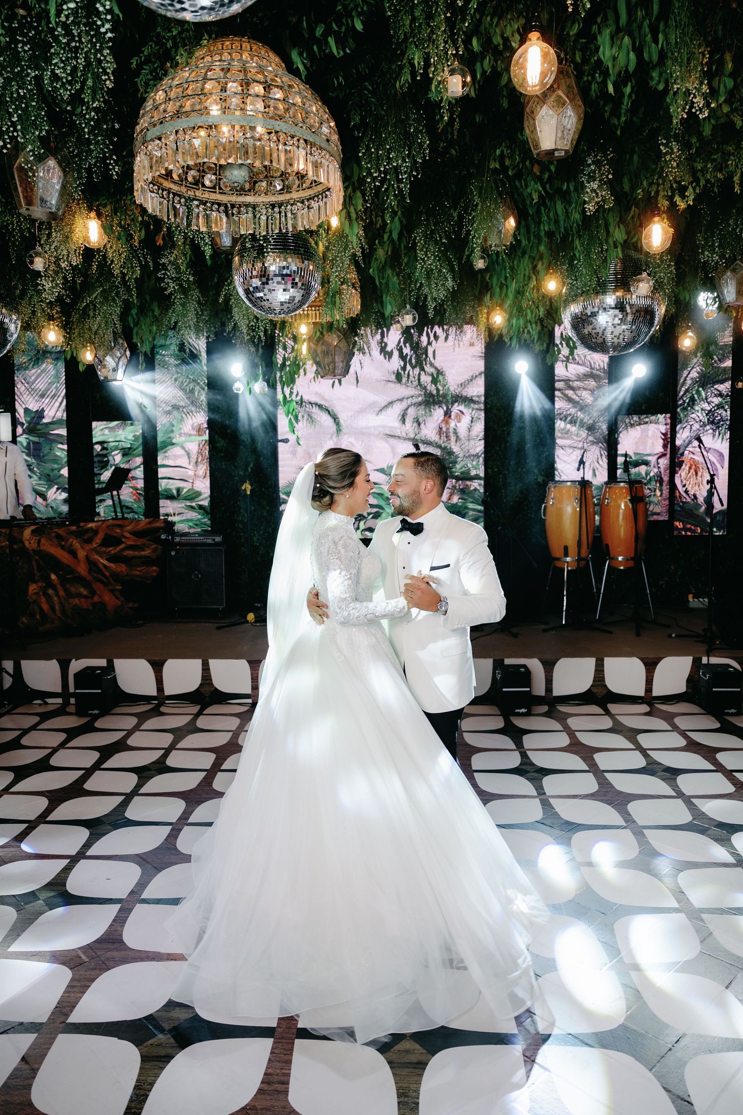 Romantic wedding couple dances together on a checkered floor under hanging lights and greenery at an elegant reception.