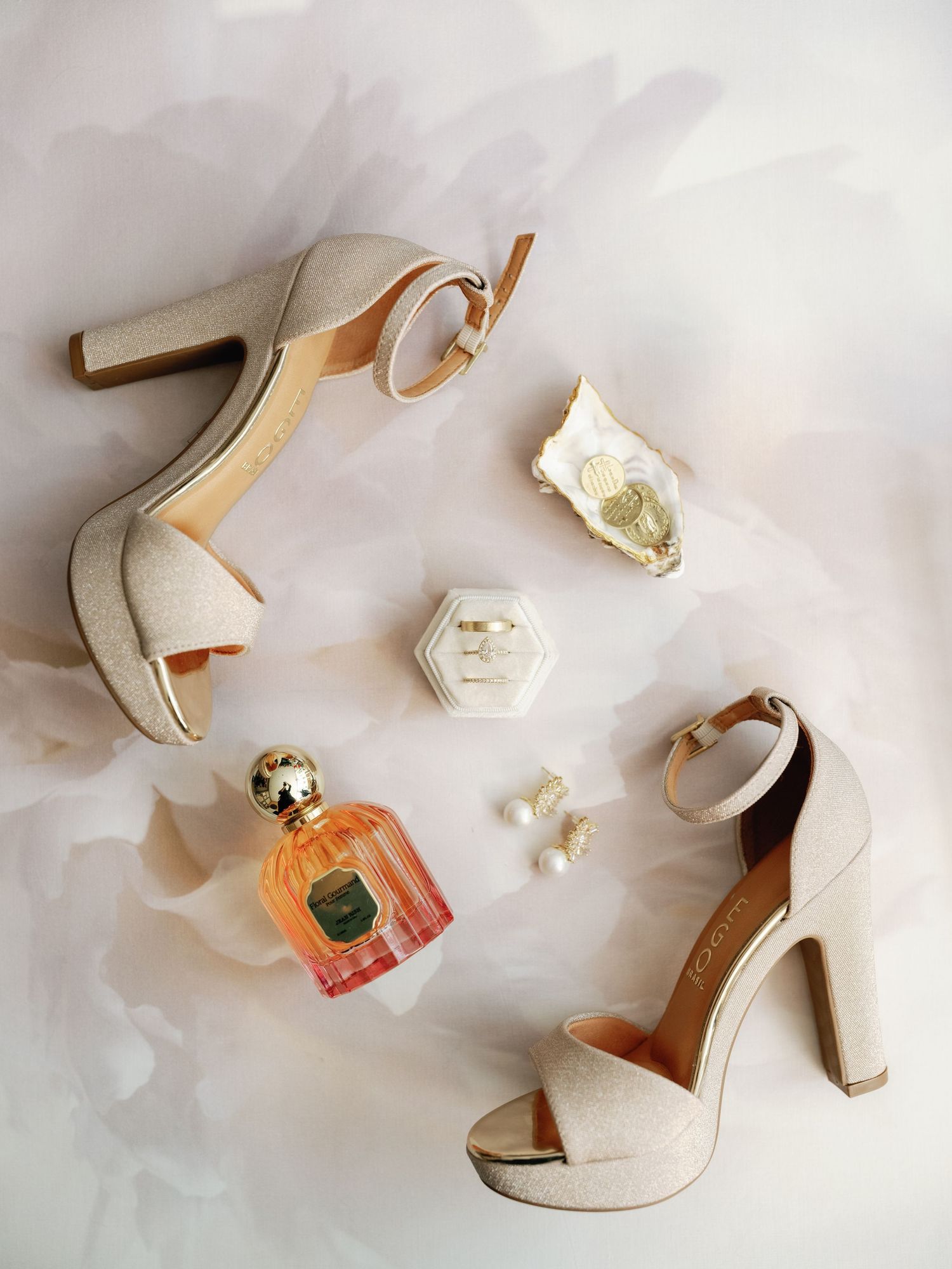 Wedding accessories including elegant beige heels, perfume bottle, and delicate jewelry arranged on light fabric.