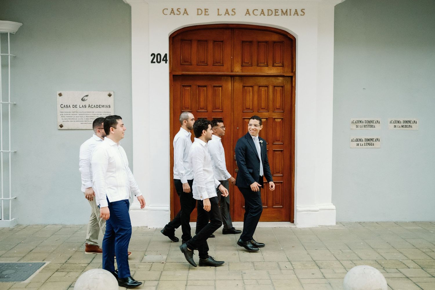 People in white uniforms walk along a white building corridor with wooden doors marked Casa de las Academias.