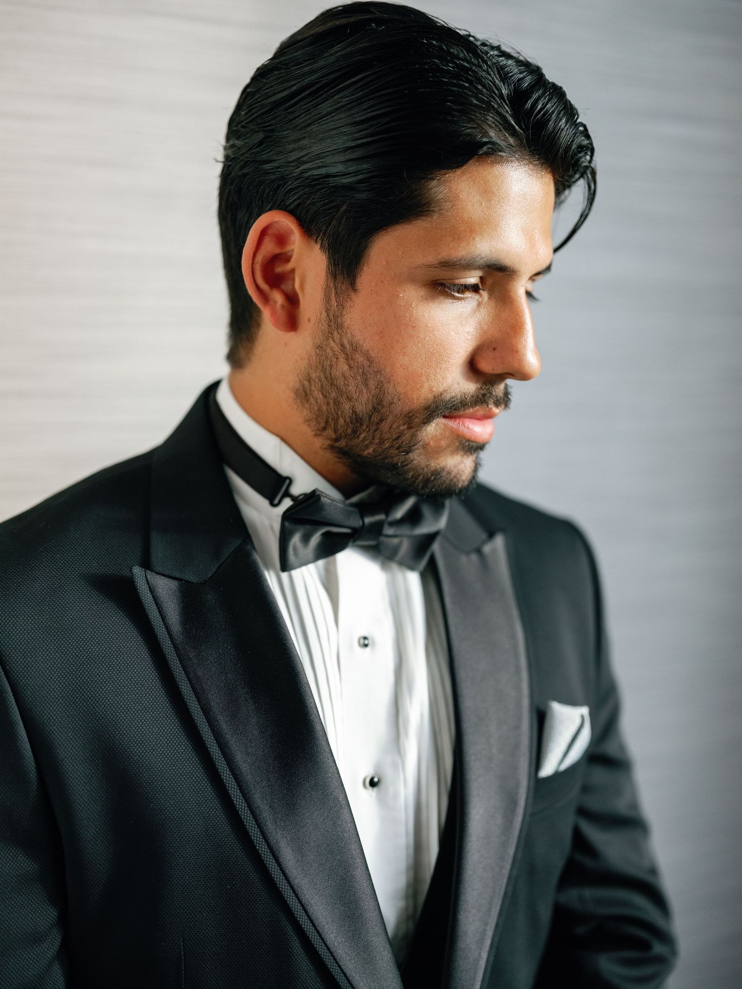 A groom in a black tuxedo and bowtie poses for a profile portrait against a light background.