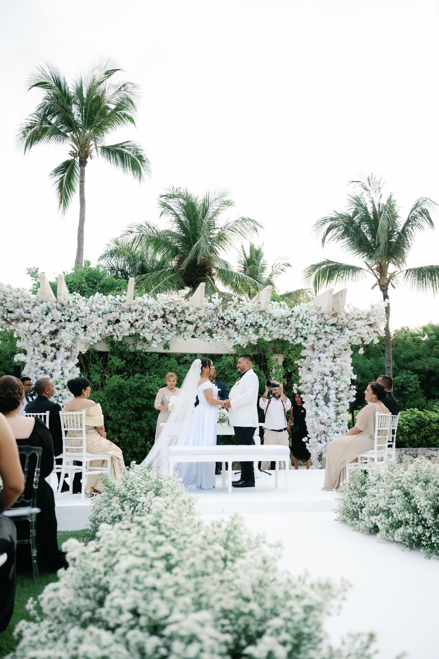 A stunning outdoor wedding ceremony setup with white floral arches and palm trees in the background.