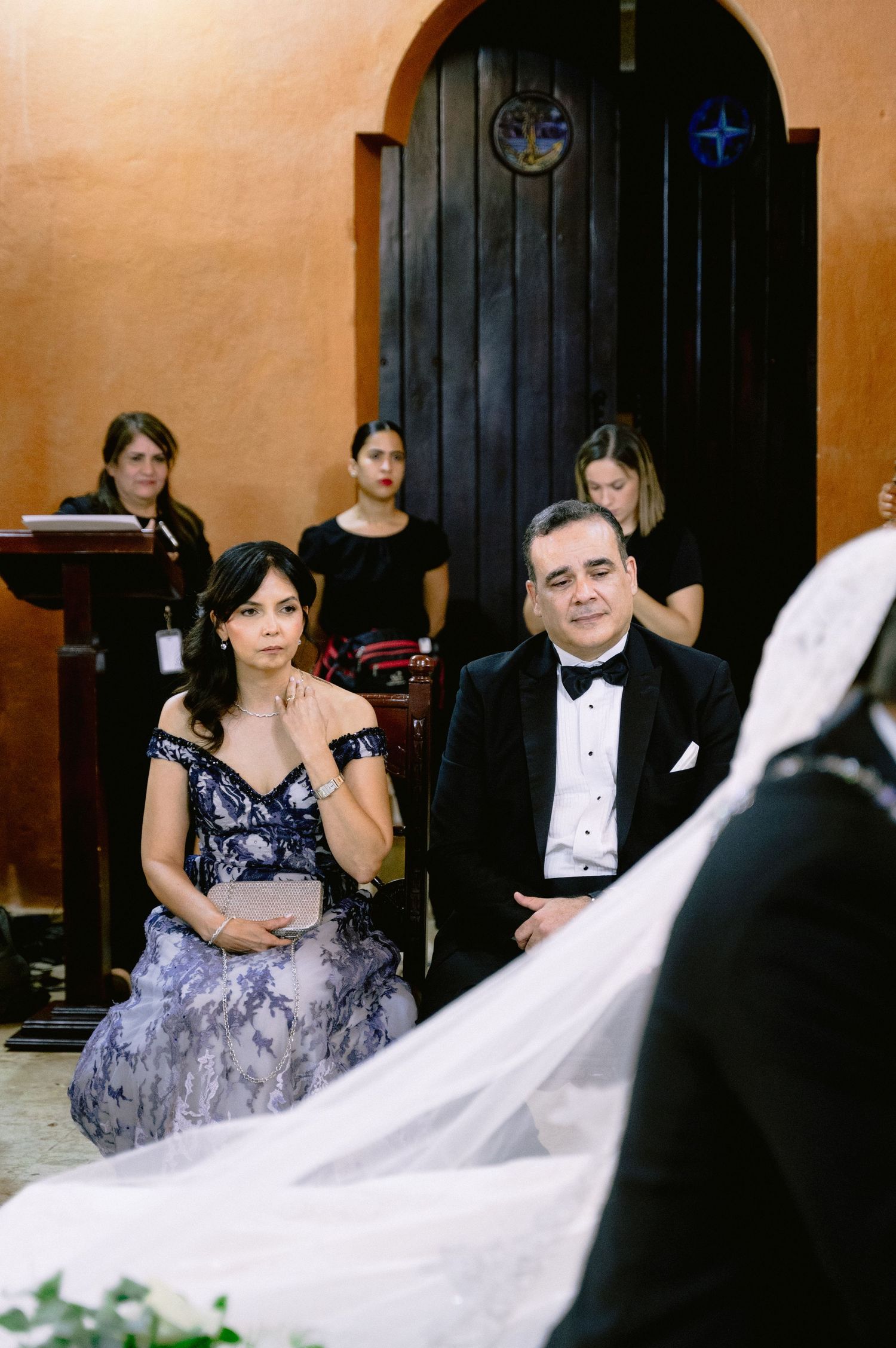 Wedding party members in formal attire seated on dark wooden chairs during indoor ceremony.