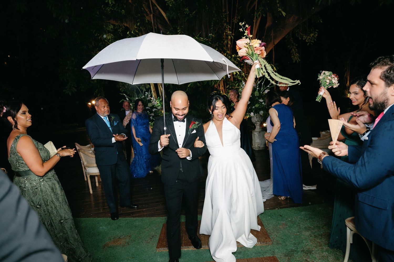 Wedding guests dance and celebrate under white umbrellas during an outdoor evening reception.