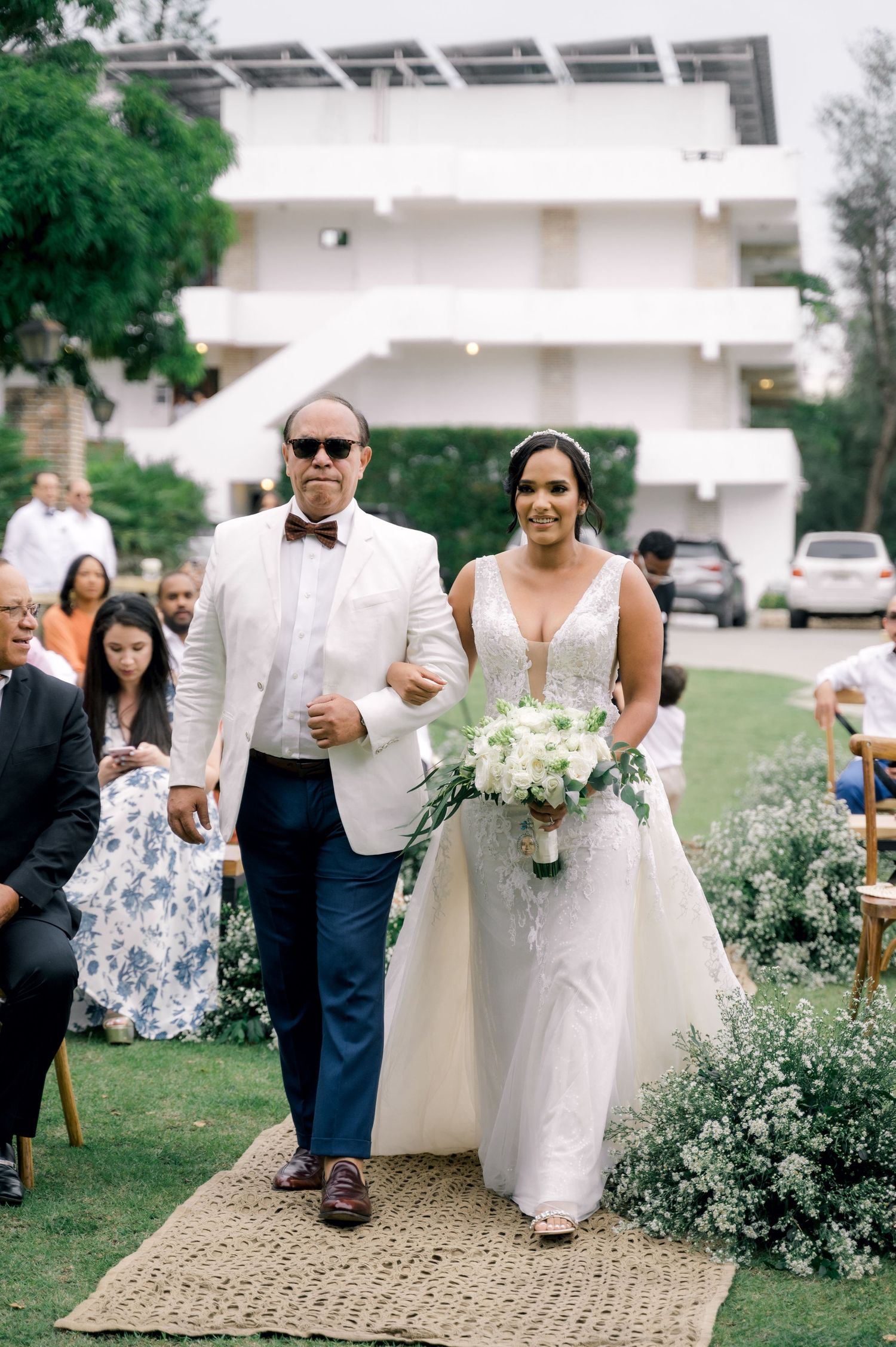 Wedding ceremony processional on an outdoor path lined with delicate white flowers and greenery.
