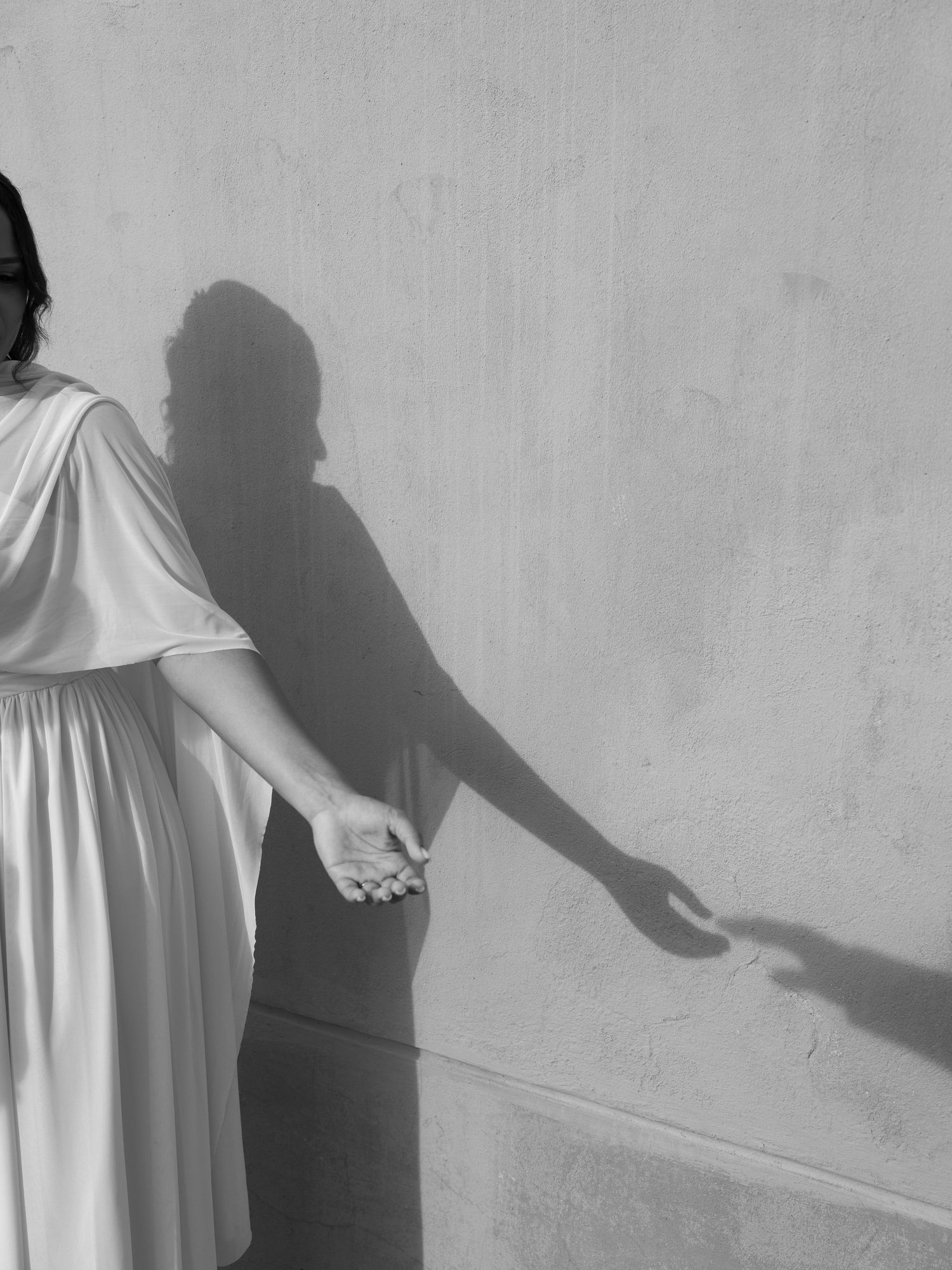 Black and white artistic photograph showing shadows cast on a wall from a figure in a flowing white dress.