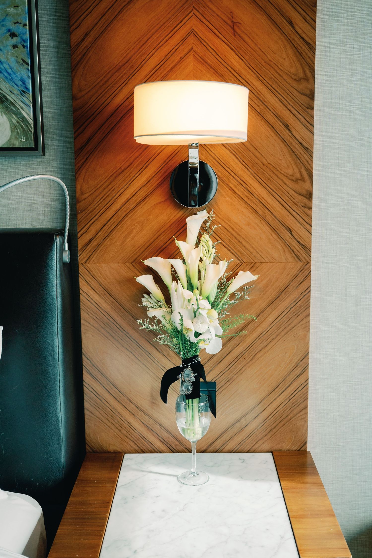 White lilies in a glass vase sit atop a marble table against a wood paneled wall with a glowing lamp overhead.
