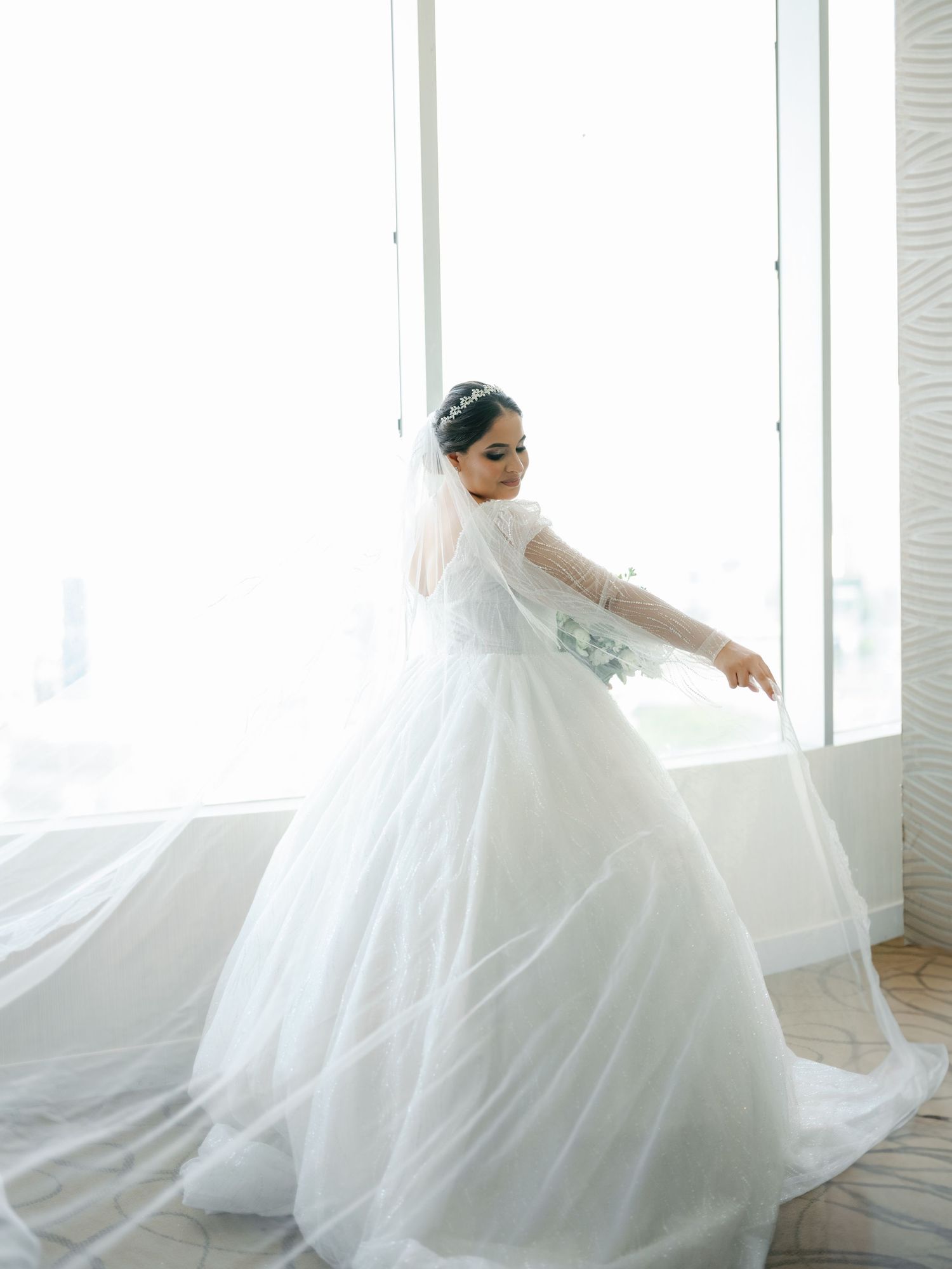 A bride in a flowing white wedding gown with long sleeves twirls gracefully by a bright window.
