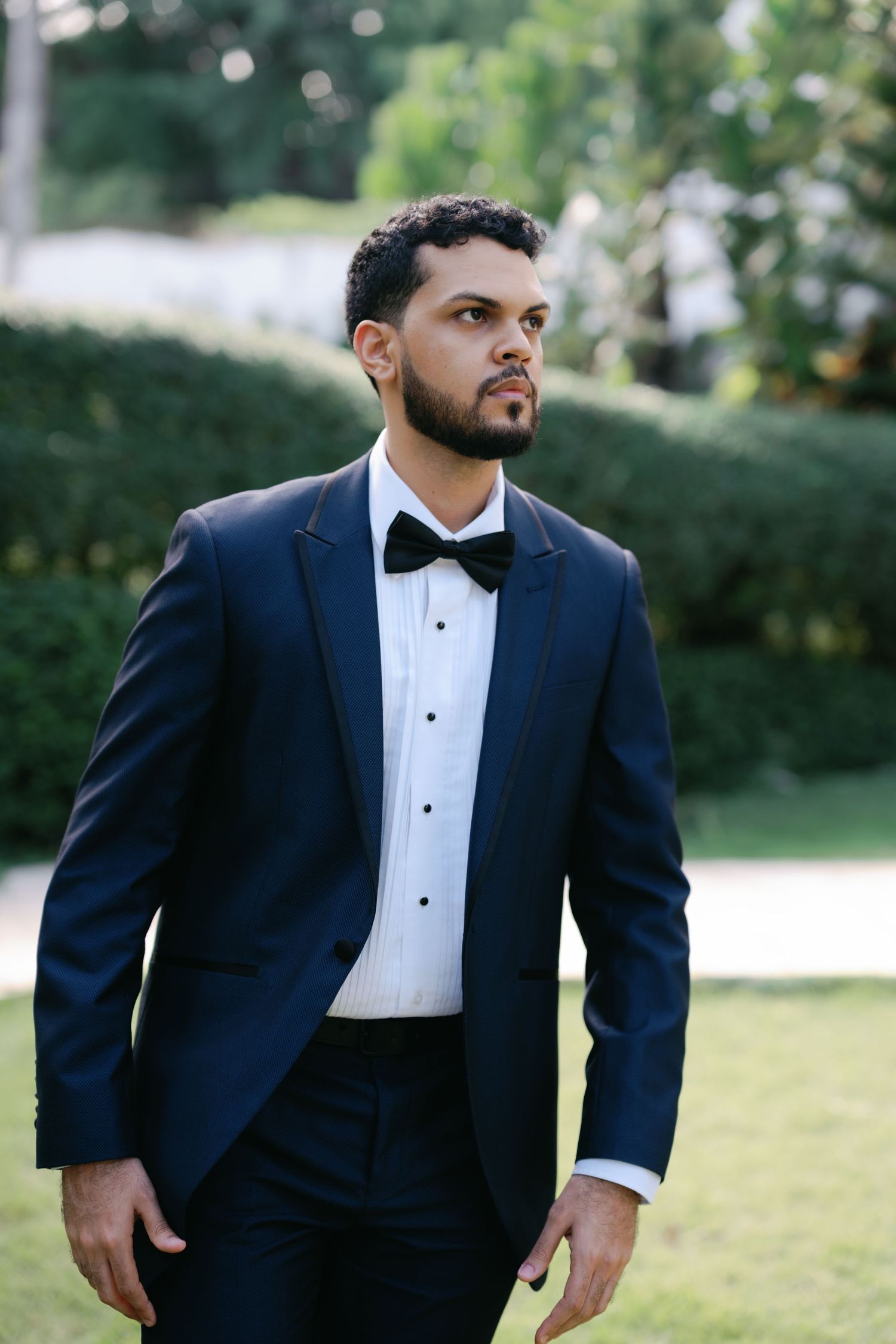 A groom in a classic navy tuxedo with black bow tie stands outdoors in a garden setting.