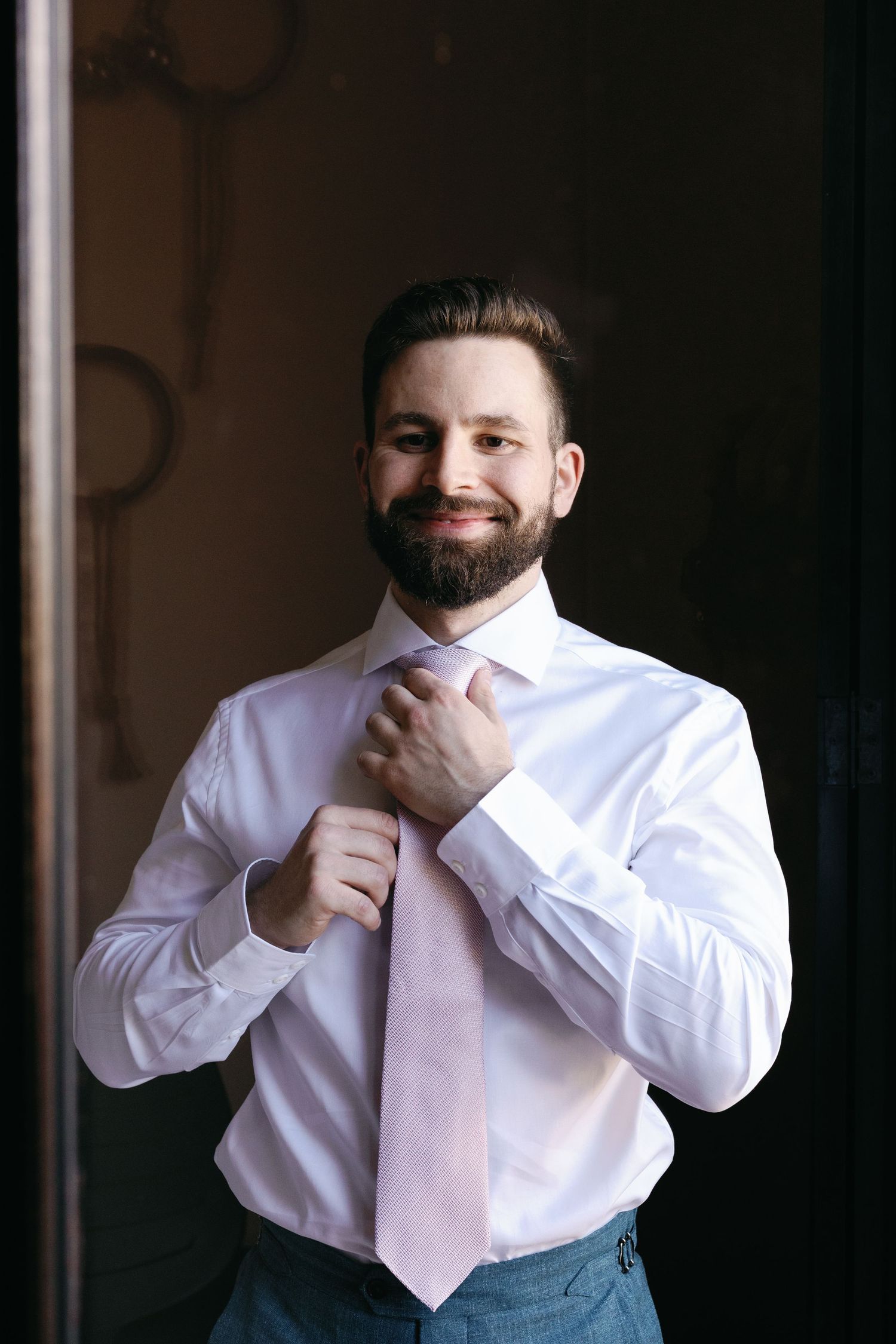 Smiling bearded person in white dress shirt and pink tie adjusts collar while standing in dark room.