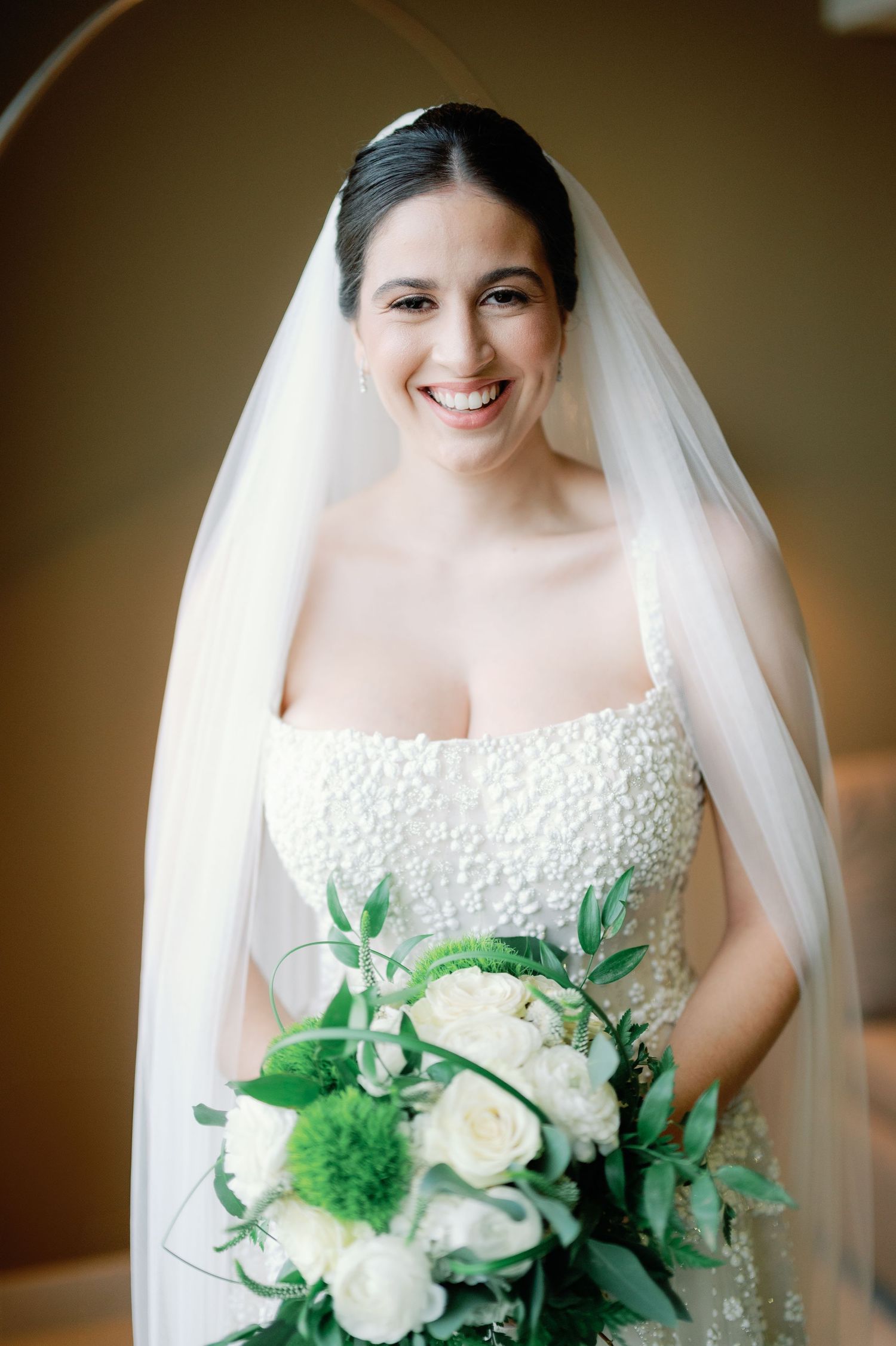 A bridal portrait featuring a strapless lace gown and white rose bouquet with greenery.