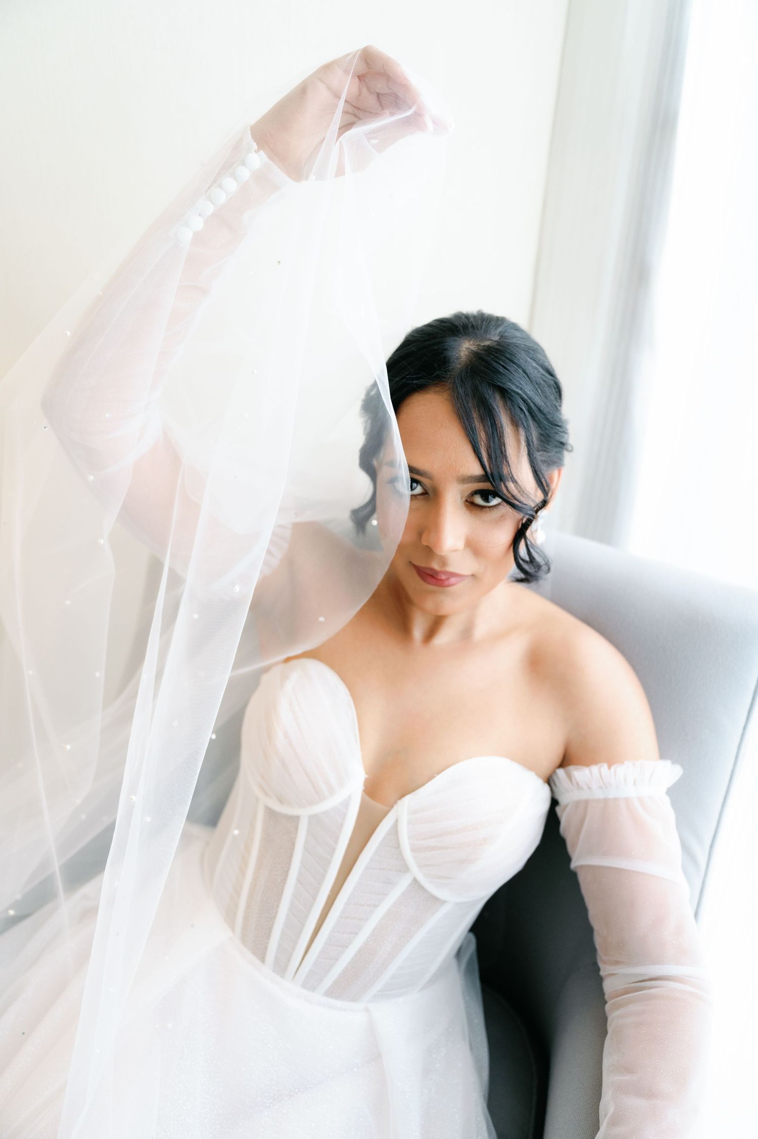 A bride wearing a white strapless gown and veil poses elegantly against a bright window background.