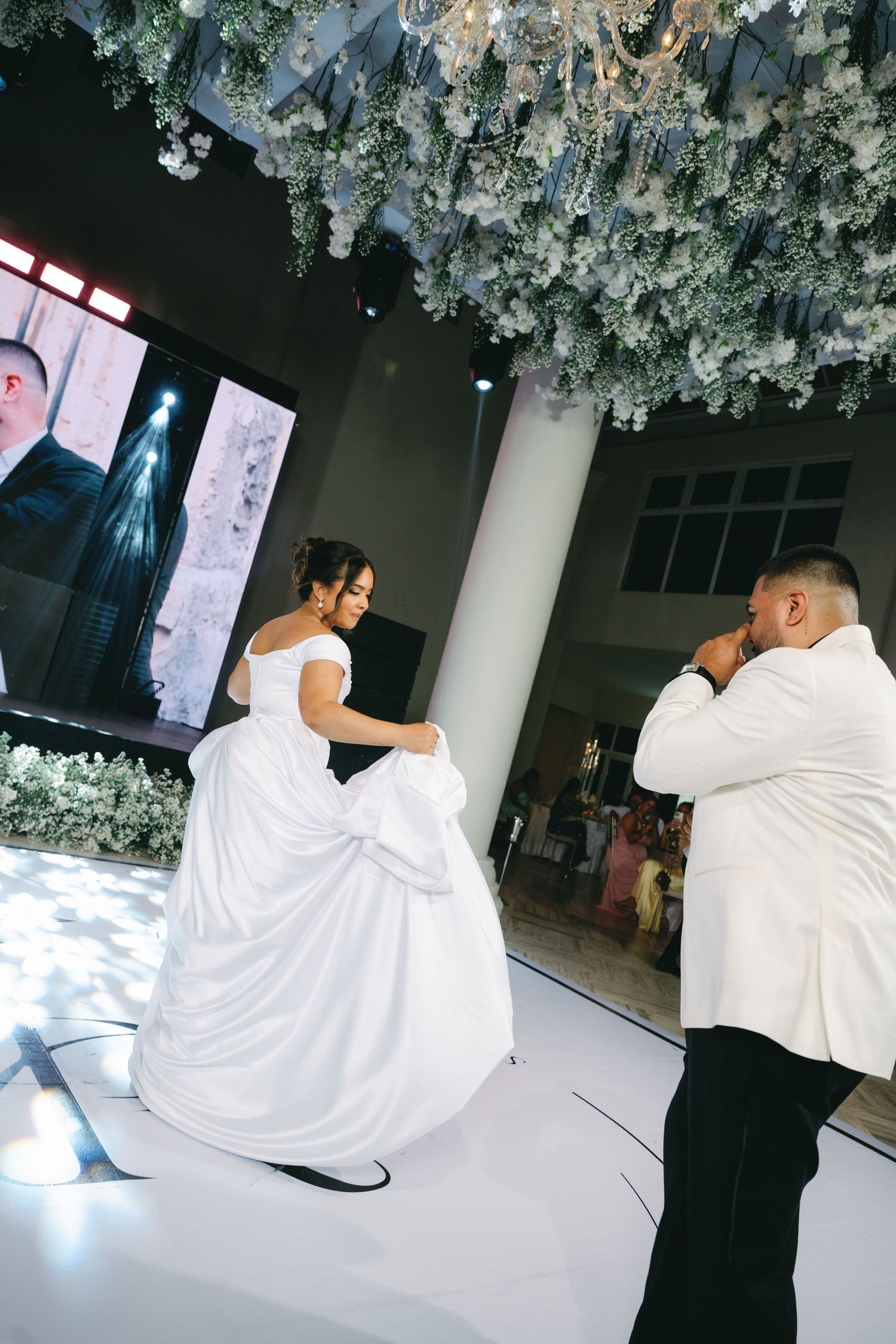 A couple performs a choreographed dance move in white attire on a reflective dance floor.