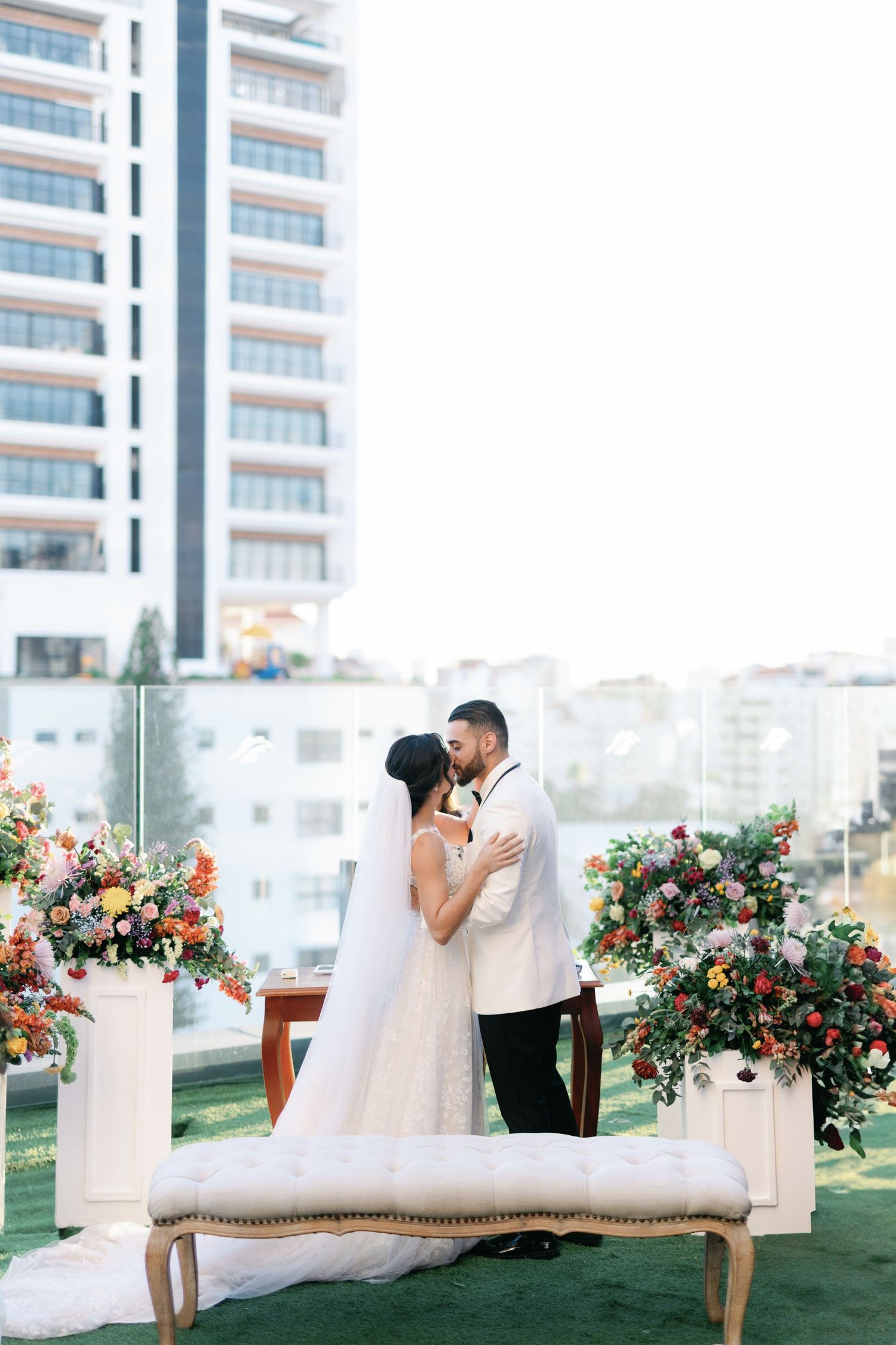 Elegant outdoor wedding setup with white chairs and vibrant floral arrangements against modern building backdrop.