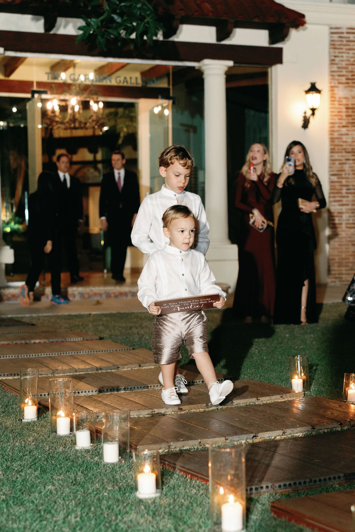 Children in white outfits play and jump around glowing candle luminaries on brick pavers at an evening event.