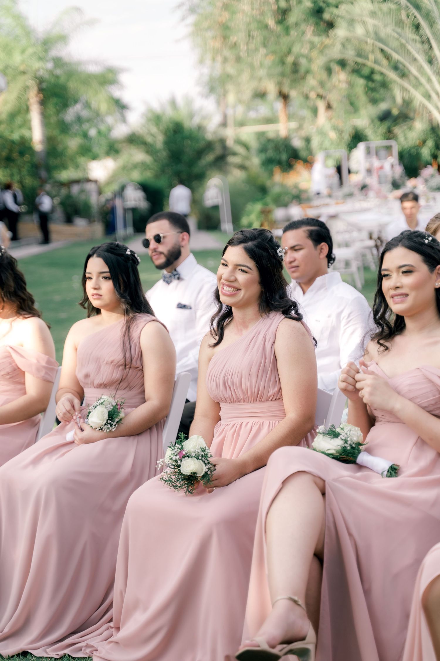 A group of bridesmaids in blush pink dresses stand together outdoors at a wedding celebration.