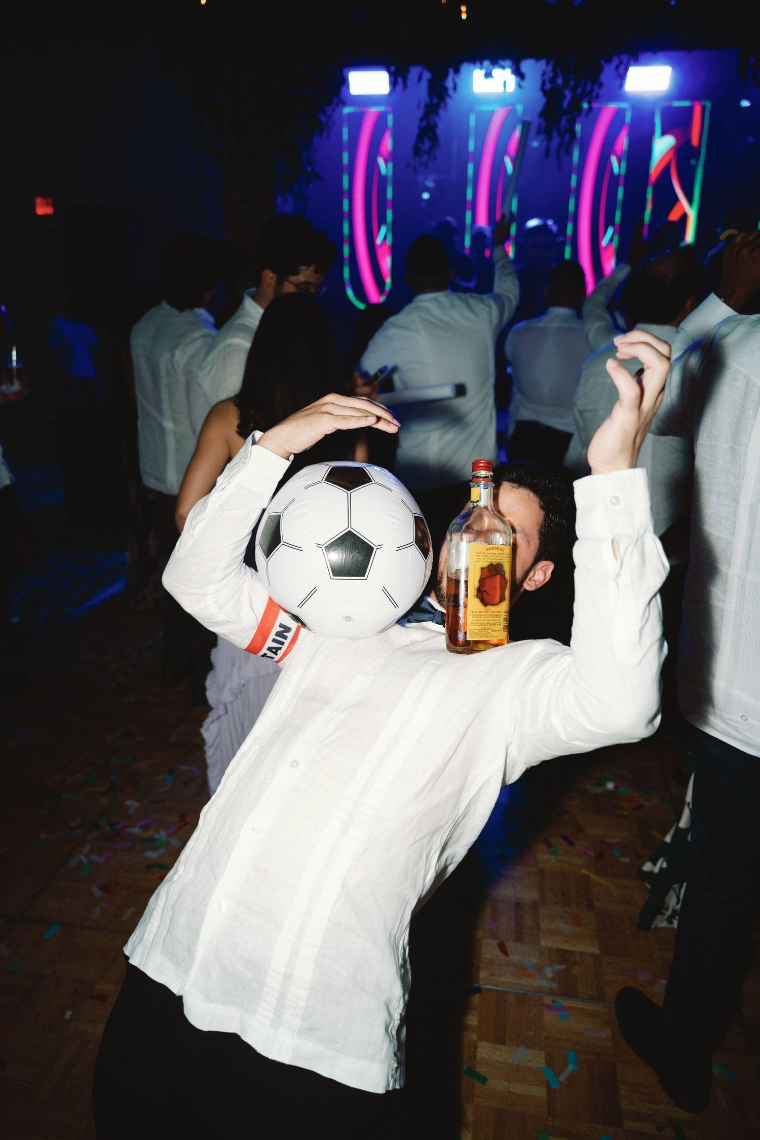 Person in white shirt wearing soccer ball mask holds up a bottle in nightclub setting.