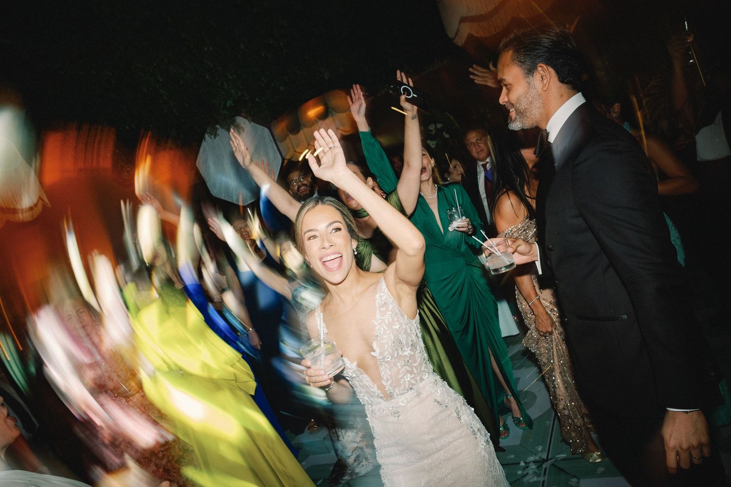 Wedding reception dance floor filled with energetic guests in motion, with colorful lights creating a festive atmosphere.