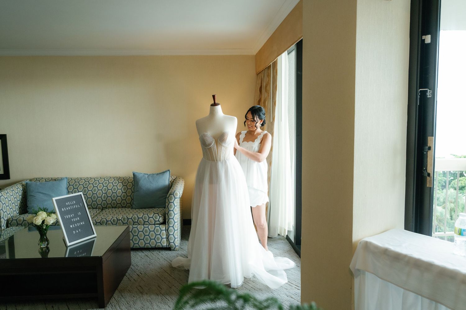 A white wedding dress on a mannequin form stands in a room with a blue couch and window.