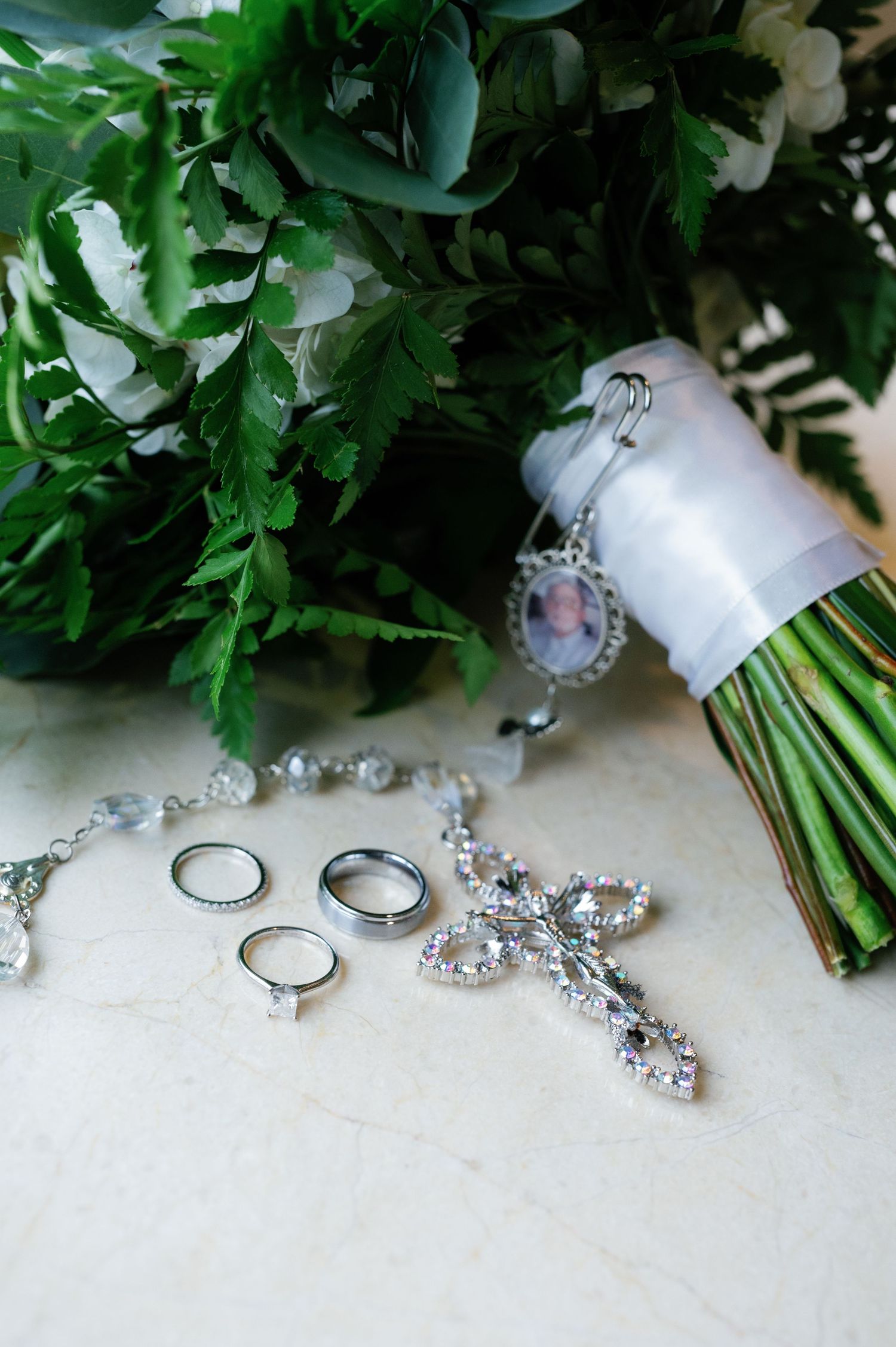 Wedding rings and crystal jewelry accessories arranged with green floral bouquet and white ribbon details.