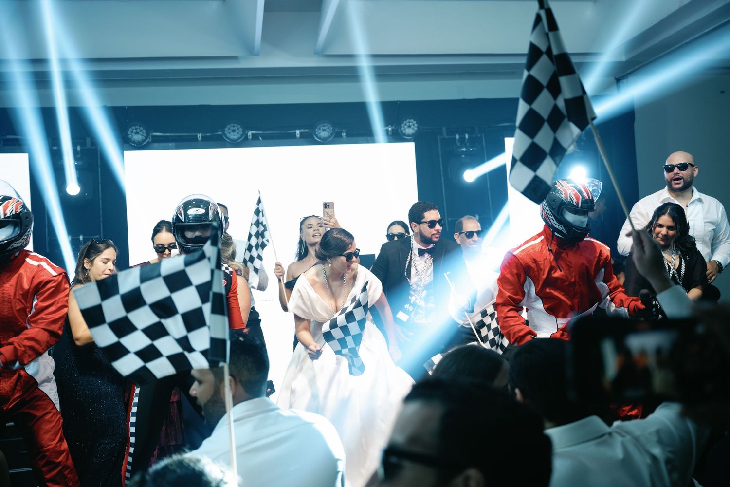 A racing crowd celebrates at a motorsport event with checkered flags and bright blue lighting effects in the background.