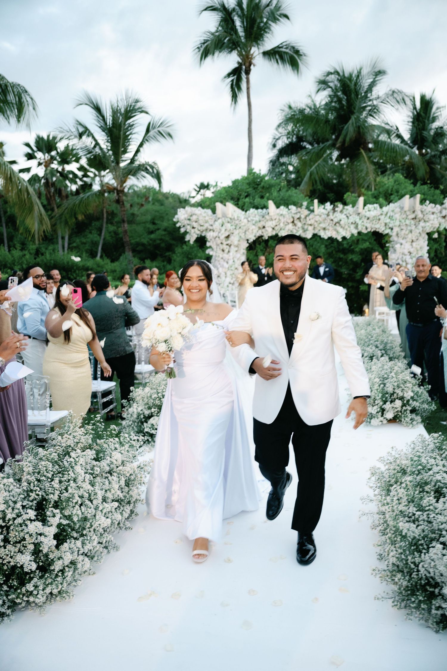 Wedding party celebrates with joyful dance moves along a tropical palm-lined path decorated with white flowers.