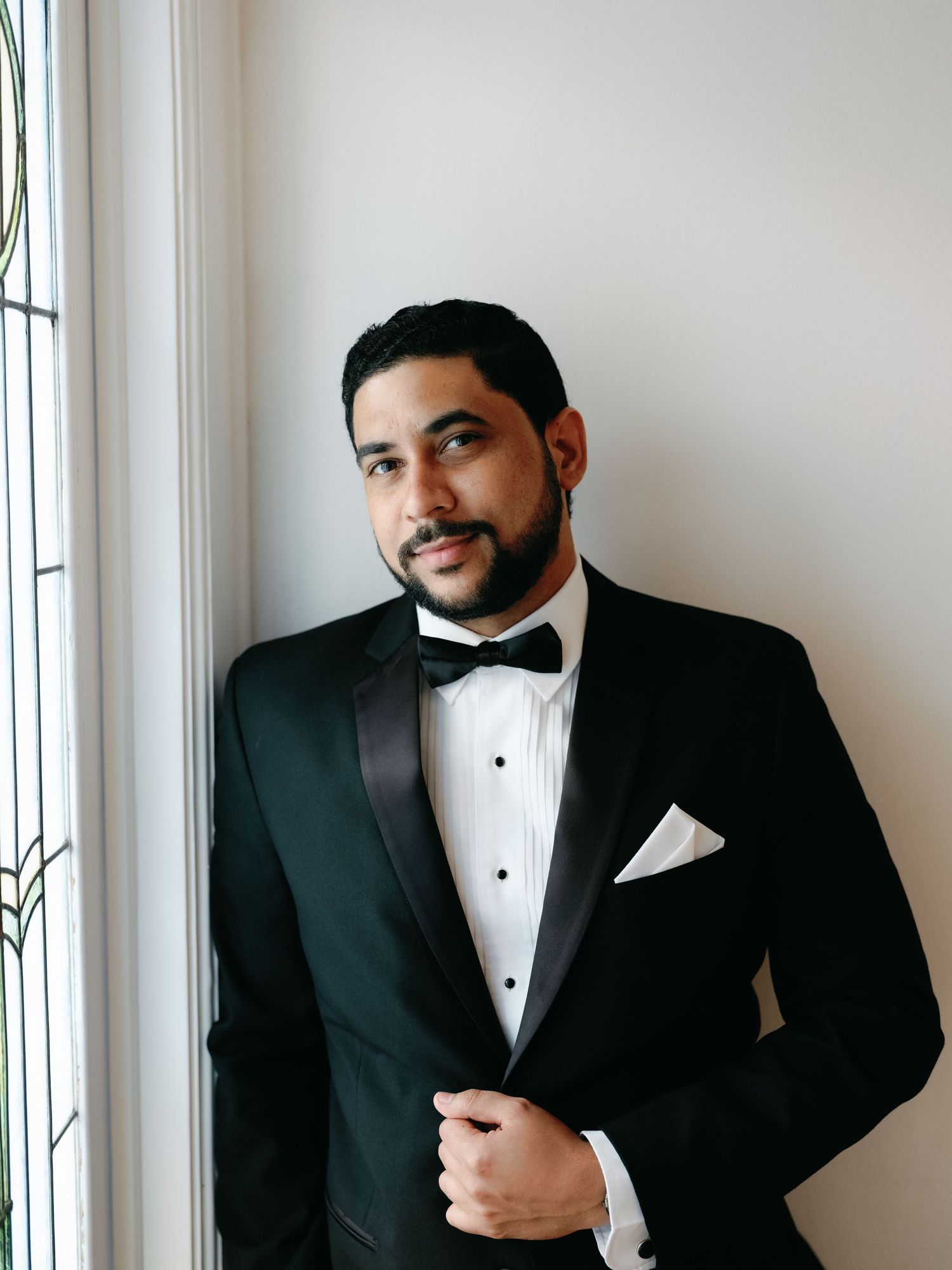 A person in a black tuxedo and bowtie poses against a white wall near windows.