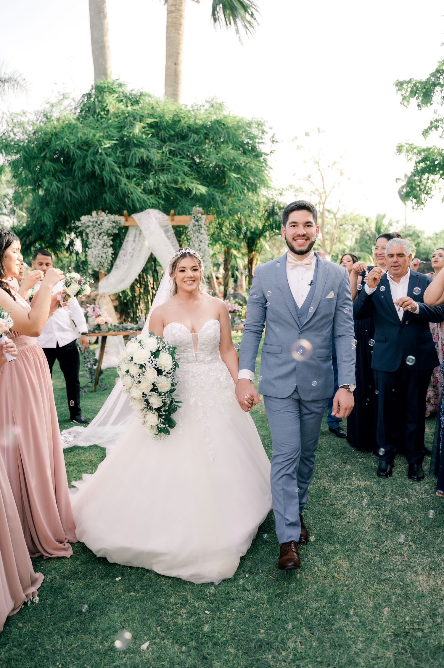 Wedding party celebrates outdoors with bridesmaids in blush pink dresses and groomsmen in gray suits.