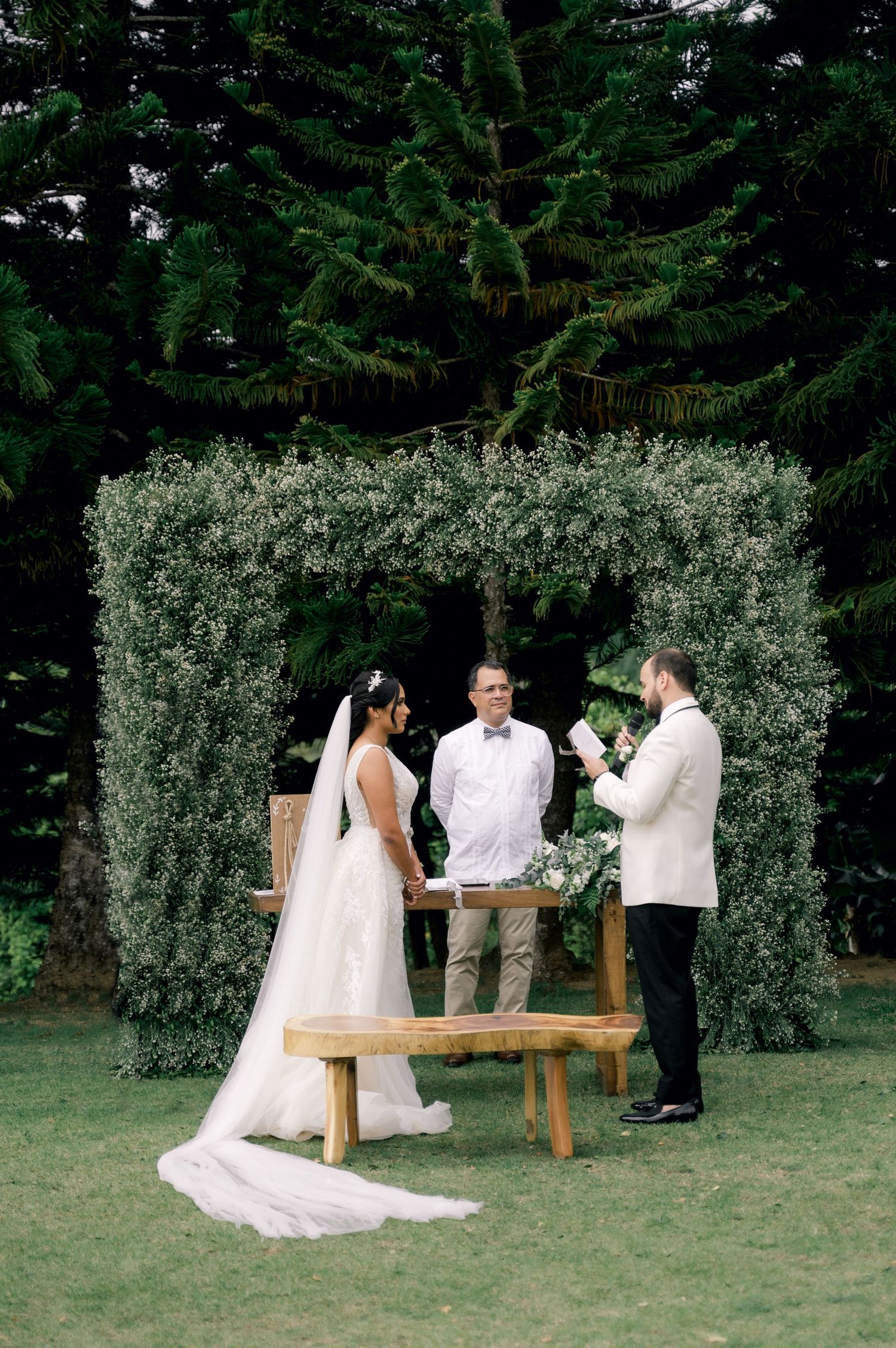 An outdoor wedding ceremony takes place under a lush greenery arch with a rustic wooden bench in a garden setting.