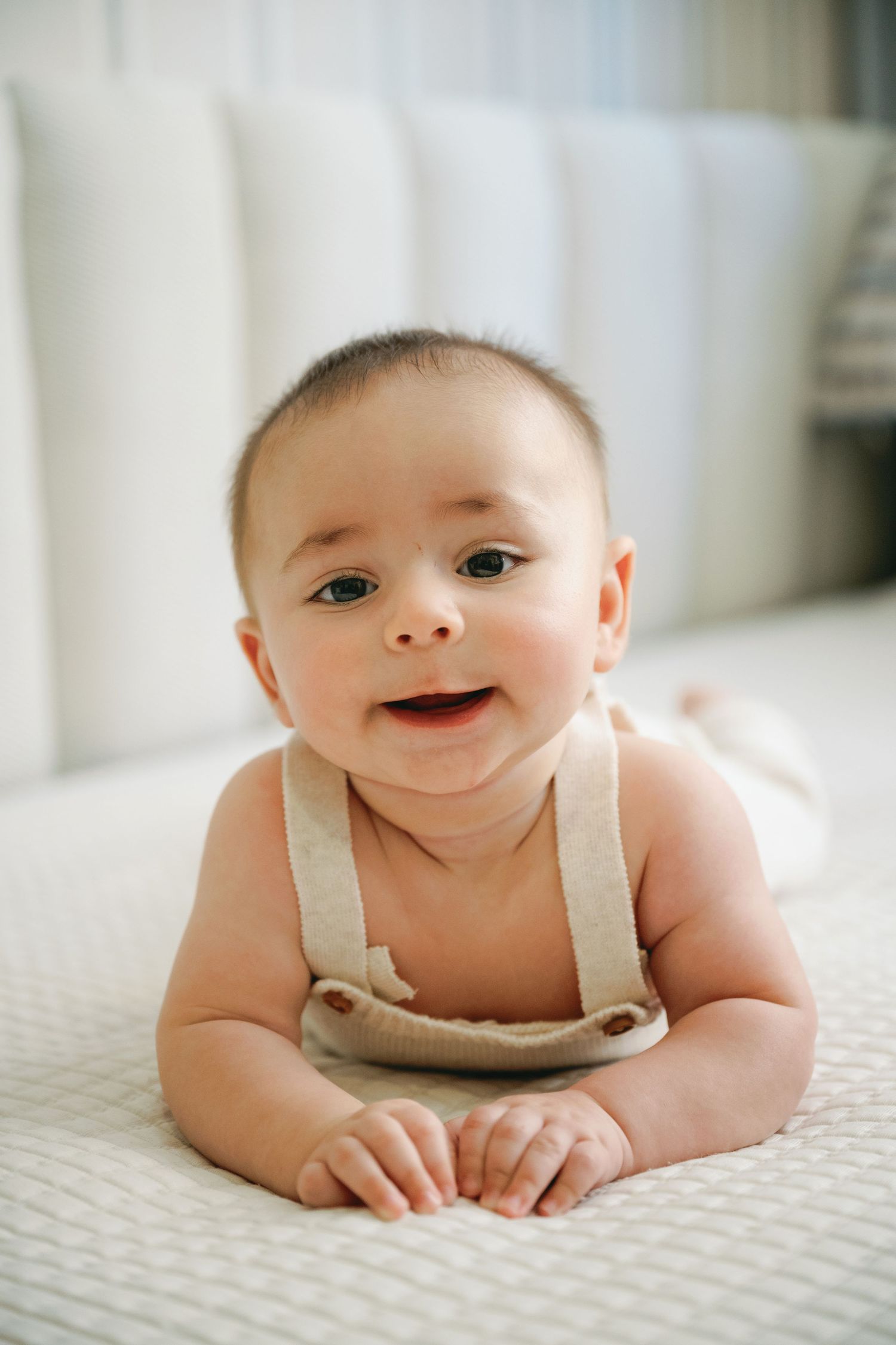 Adorable baby in linen suspenders doing tummy time on a white textured surface, smiling at the camera.