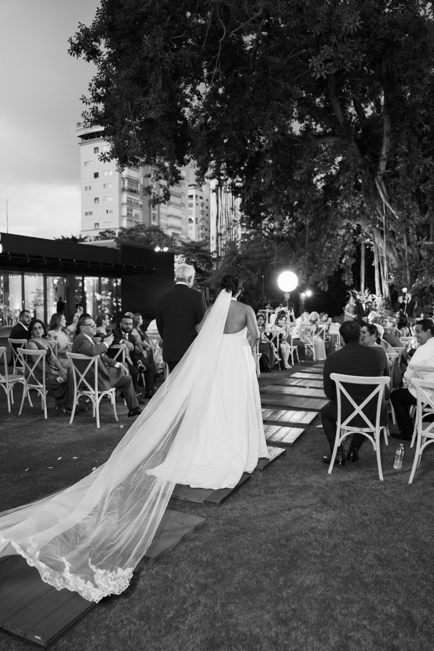 A flowing wedding dress train trails across a grassy lawn during an outdoor reception with chairs and guests visible.