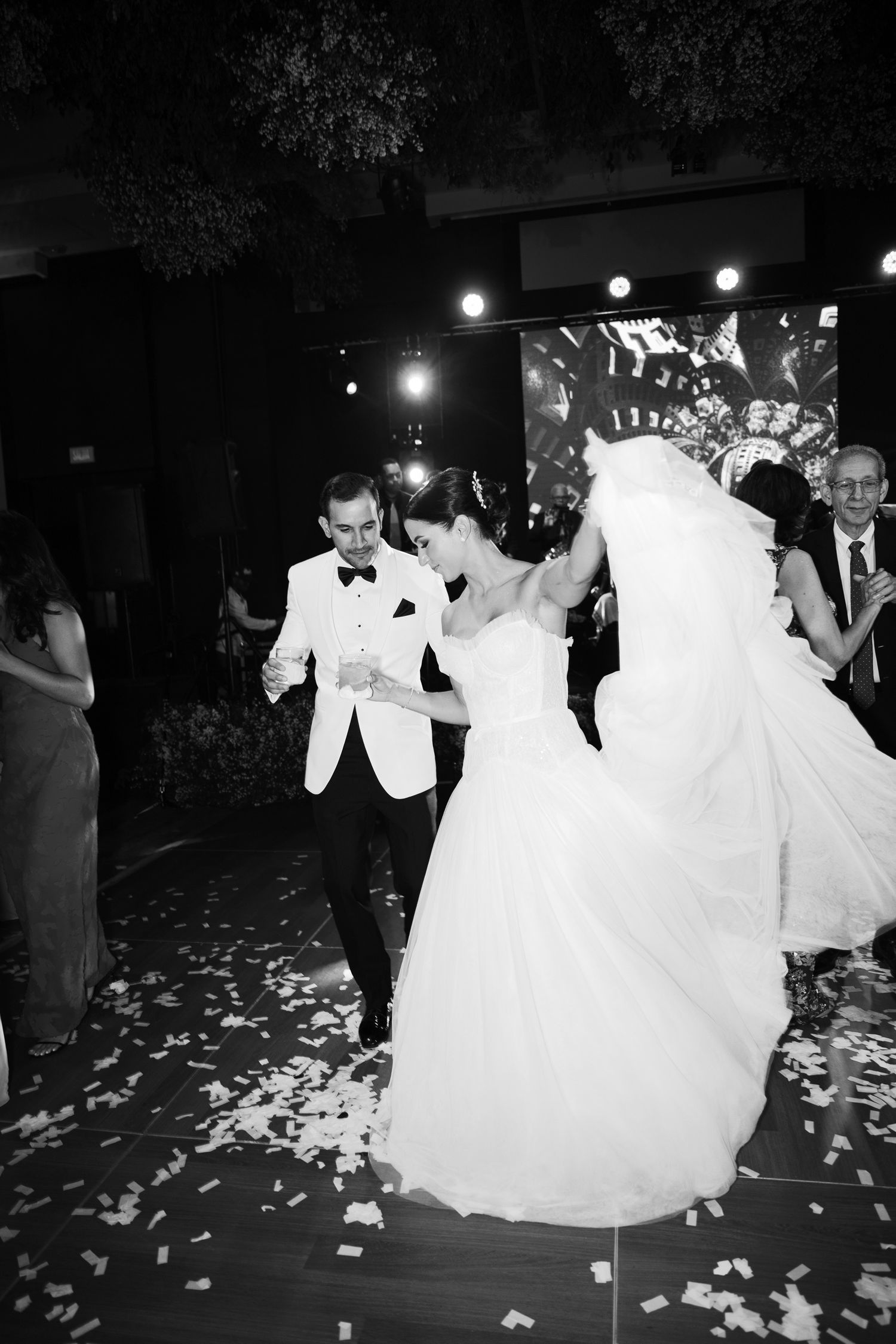 A couple dances together at their wedding reception while rose petals scatter across the dark dance floor.