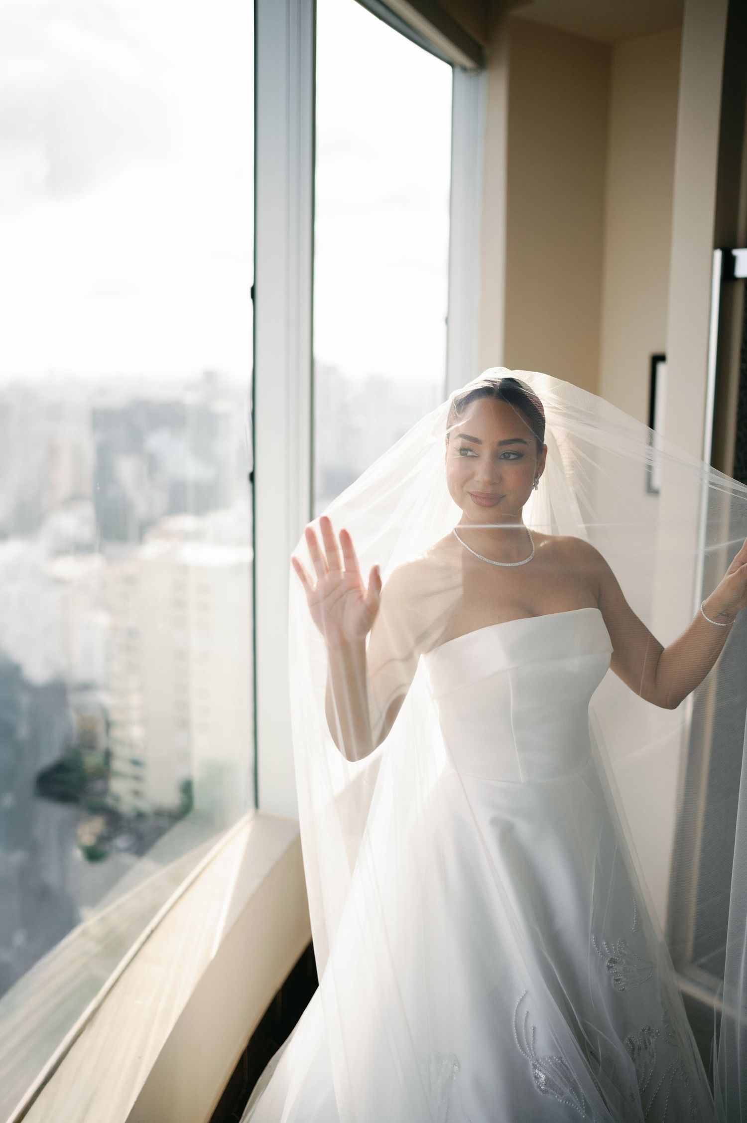 A bride in a white wedding dress and veil stands by a window in a series of ethereal poses with soft natural light.