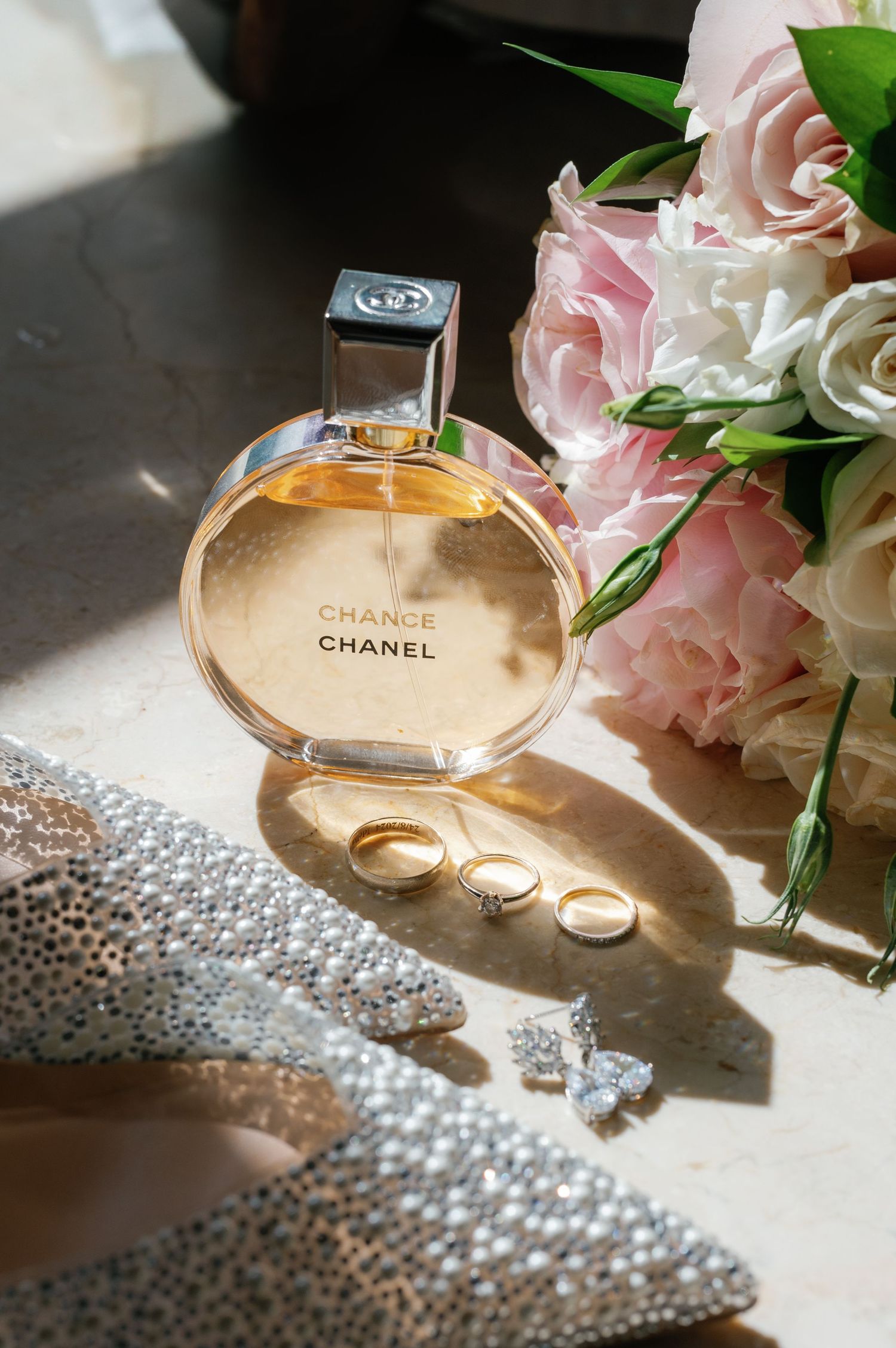 Chanel perfume bottle and wedding rings styled with pink roses and pearl beads in elegant wedding detail photography.