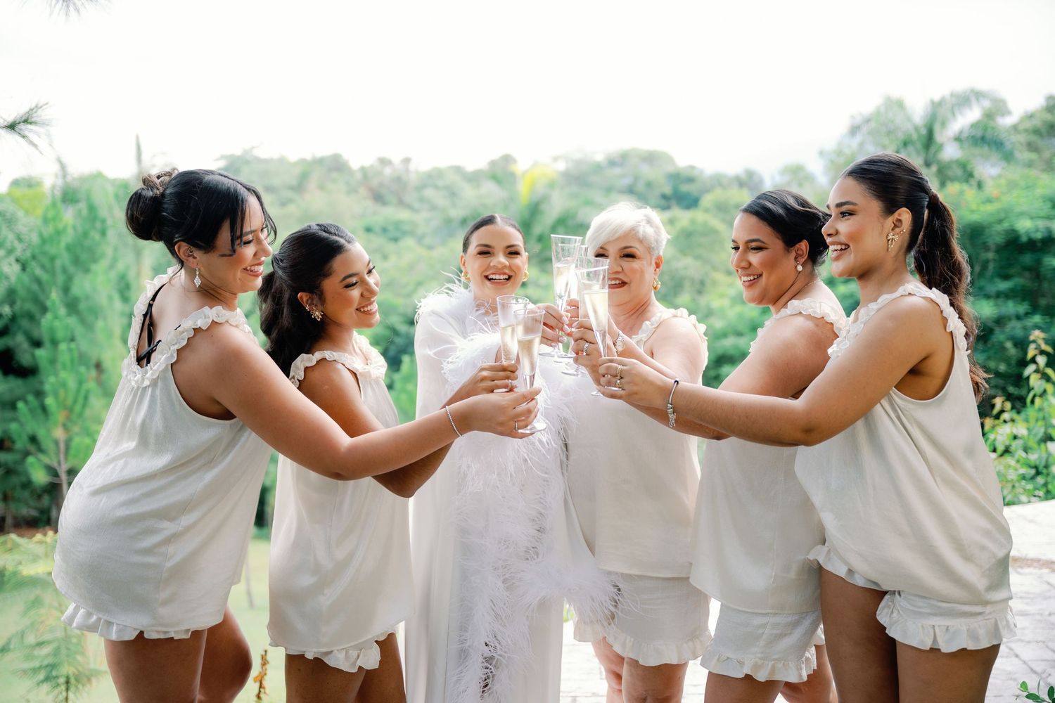 A group of people wearing white outfits laugh and celebrate together in a lush outdoor garden setting.