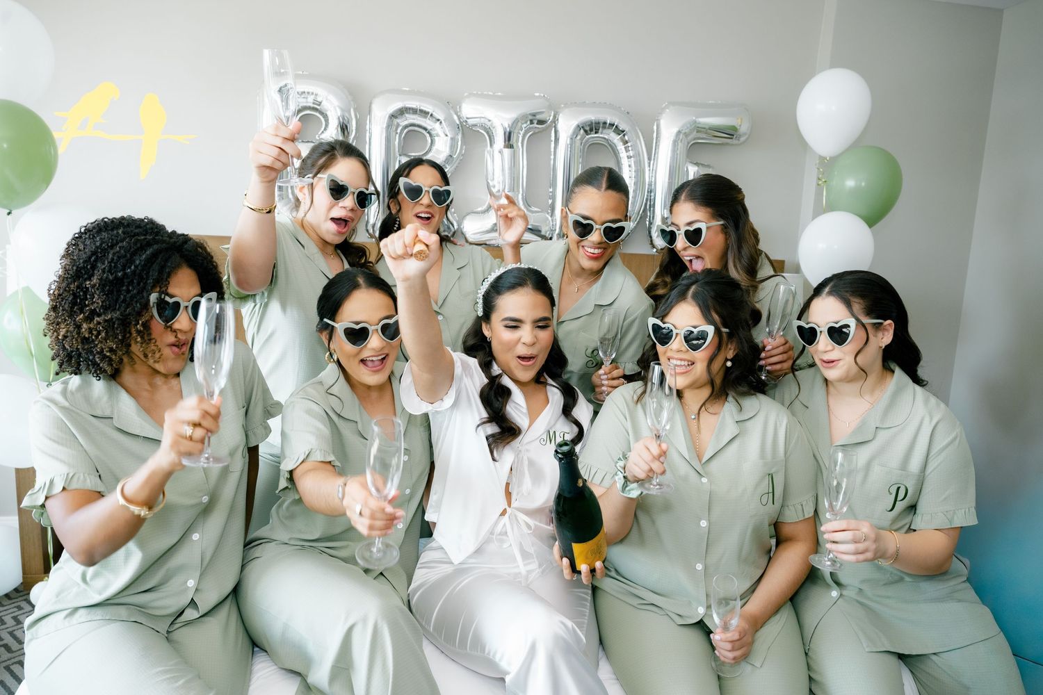 Group wearing sunglasses and matching gray outfits pose together with white balloons in bright indoor setting.