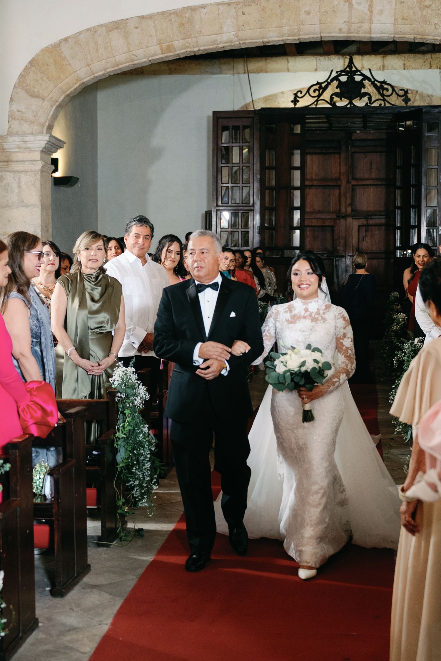 Wedding party members walk down red carpet aisle during indoor ceremony at elegant stone venue.