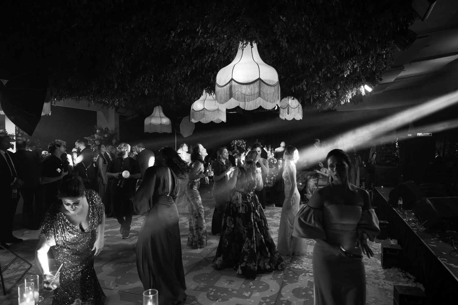 Black and white photograph of people dancing under vintage pendant lamps at a nighttime social gathering.