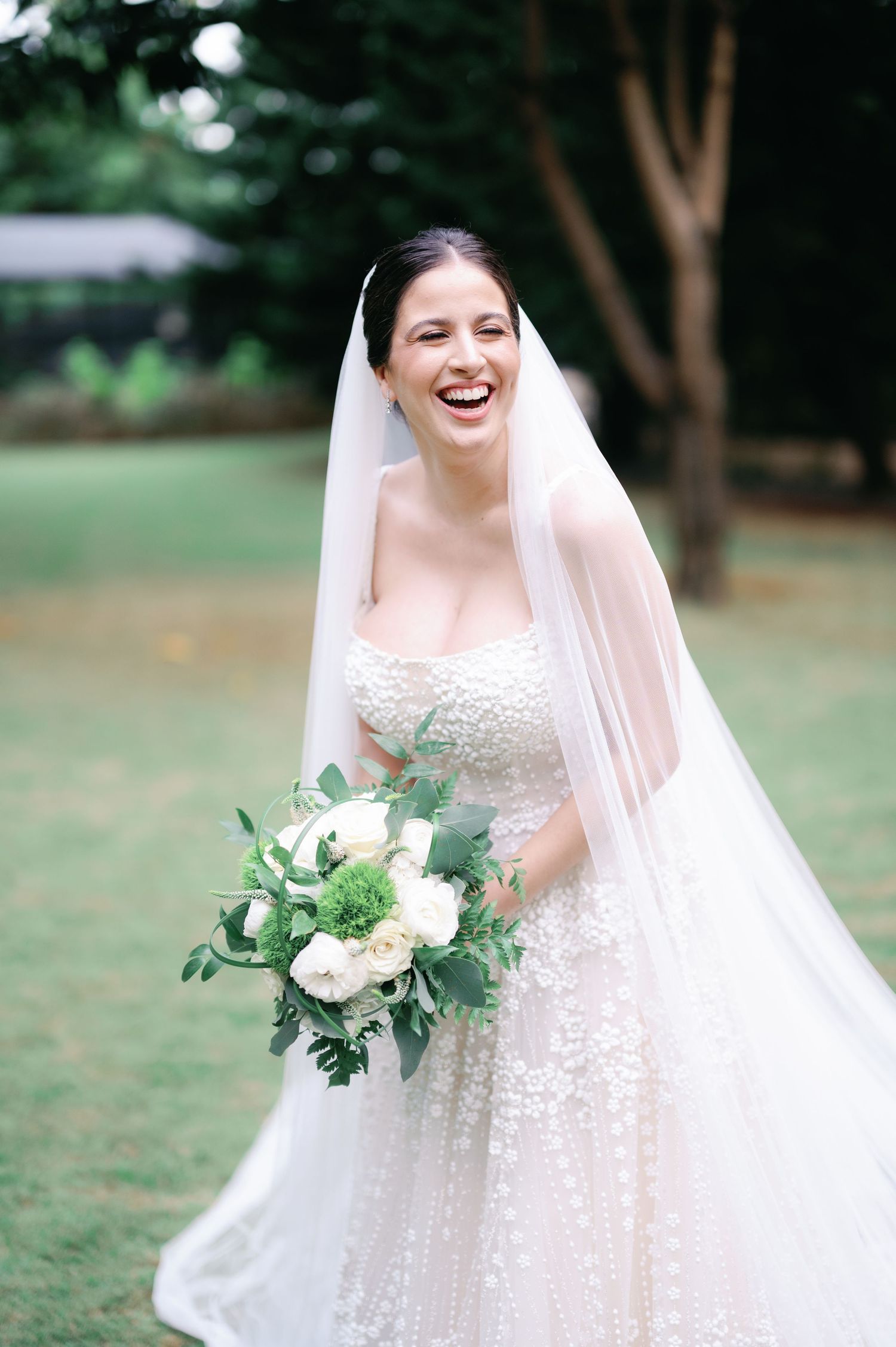 A joyful bride in a white lace gown holds a green and white bouquet while standing outdoors.