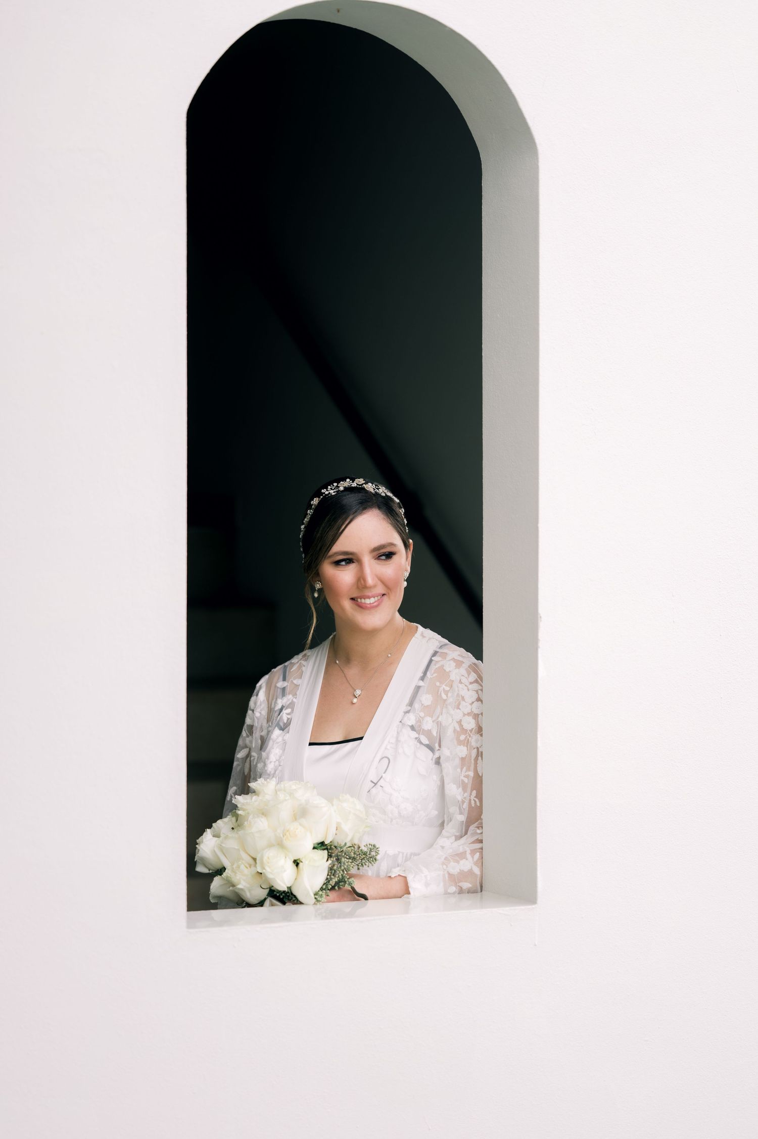A bride wearing a lace overlay dress poses in an arched window alcove with white roses.