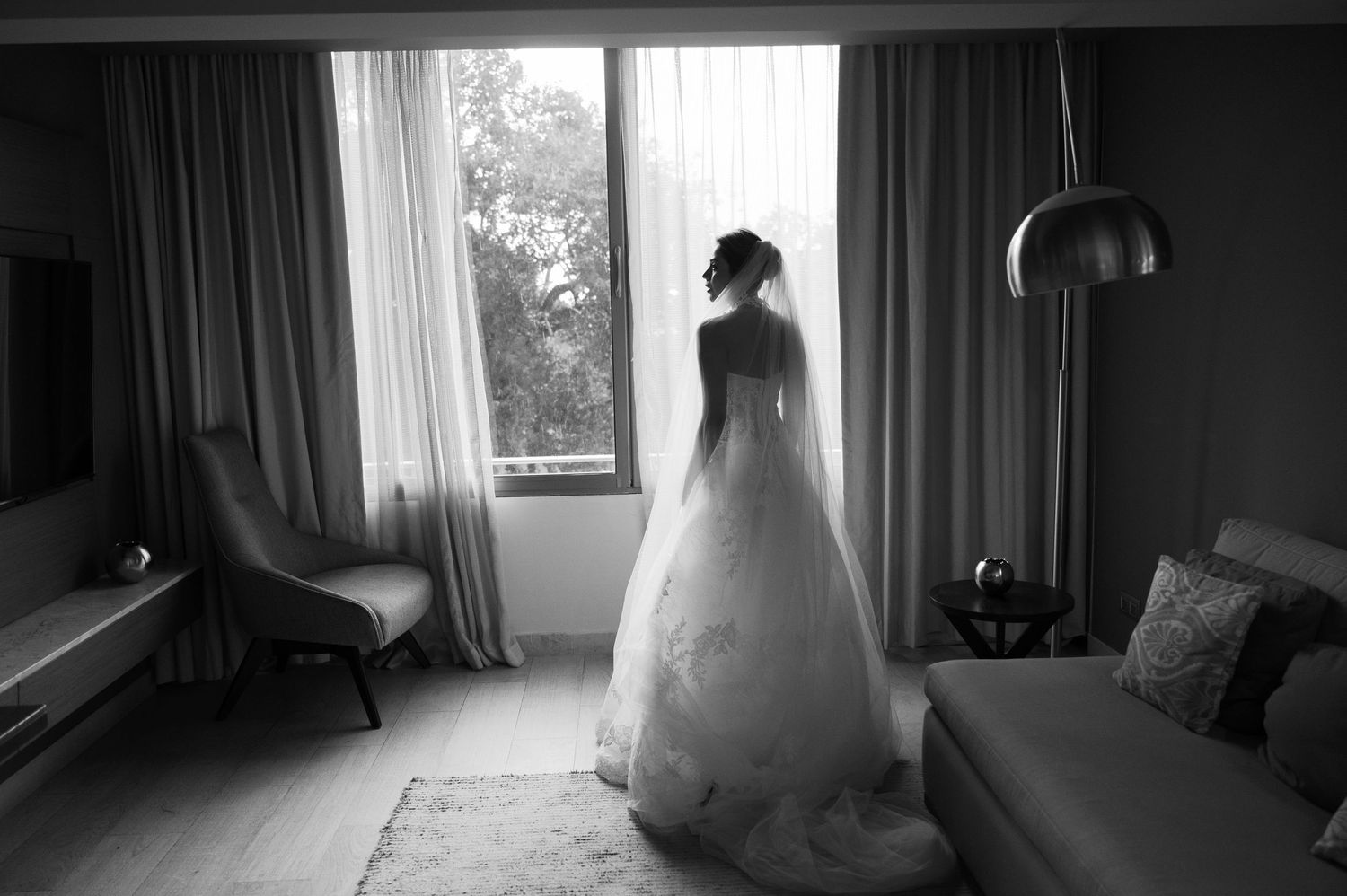 A silhouetted figure in a wedding dress stands by a window in a modern hotel room.