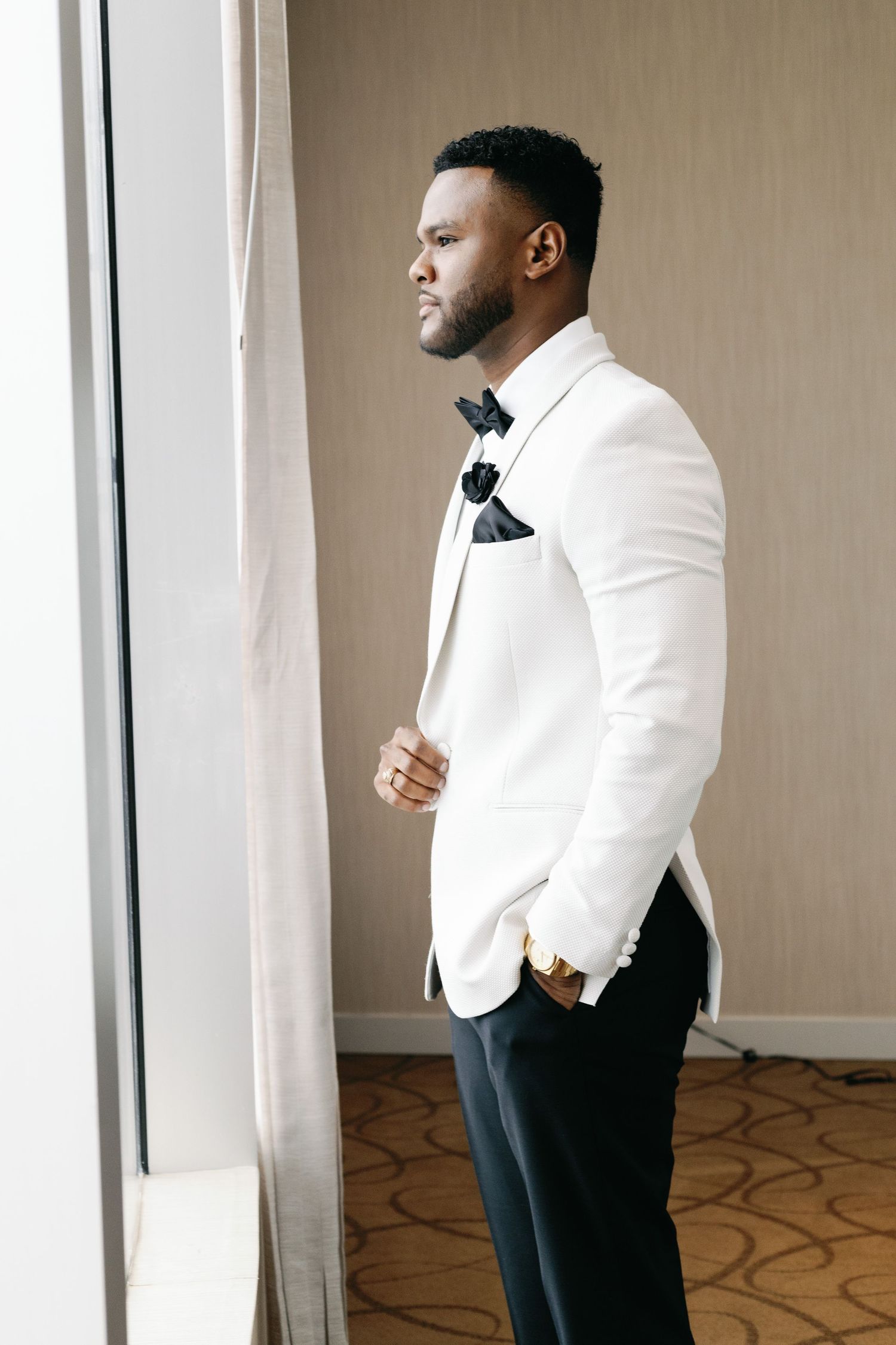 A groom in a crisp white tuxedo jacket and black bow tie stands thoughtfully by a window.
