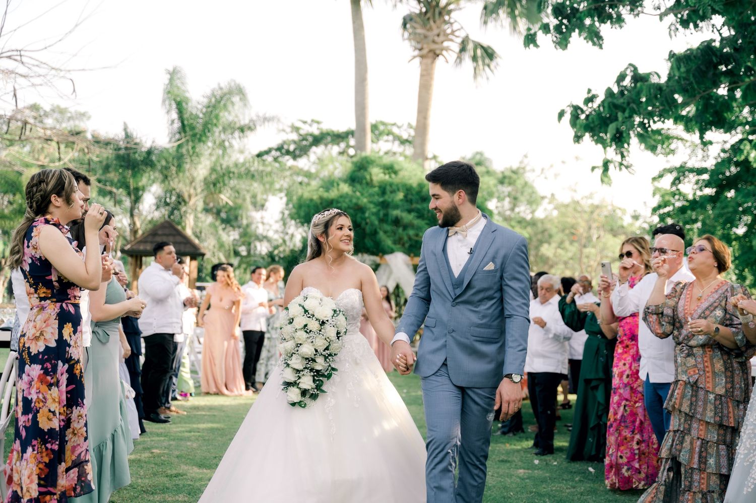 A joyful wedding ceremony takes place outdoors with guests lining the aisle amid lush tropical palm trees and greenery.