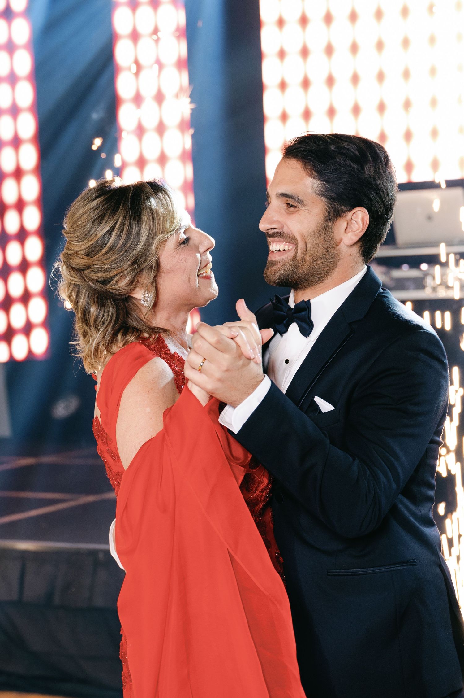 Couple in formal attire dancing and laughing together against illuminated red and white wall decor.