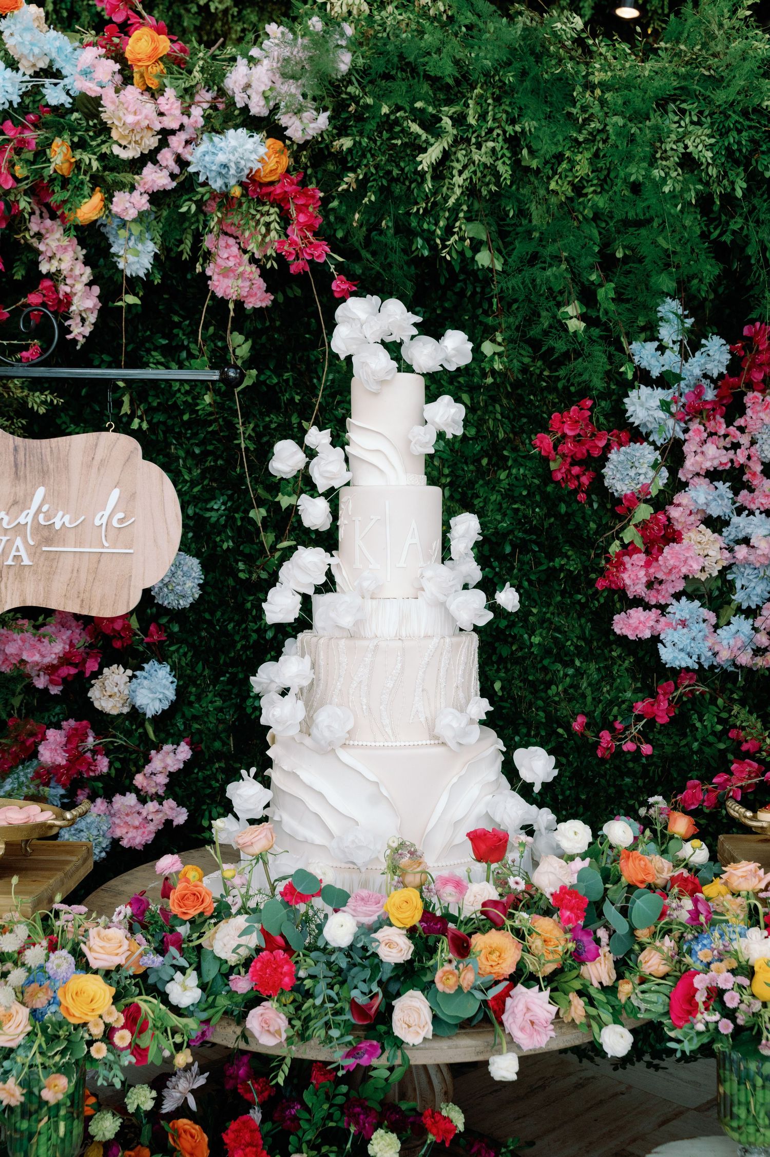 Elegant white wedding cake surrounded by colorful roses and floral arrangements.