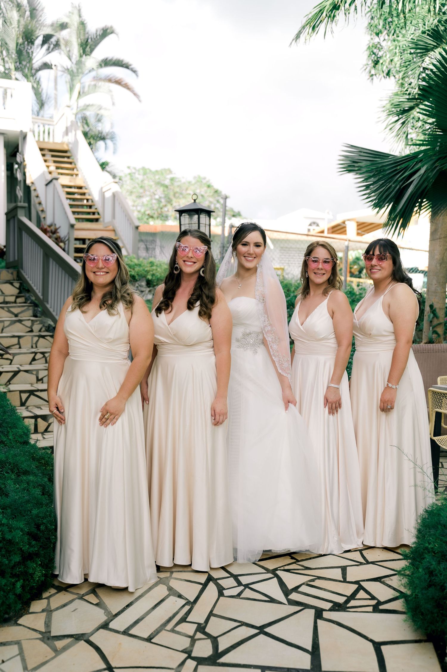 A bridal party wearing champagne-colored dresses poses together on an outdoor terrace with palm trees.