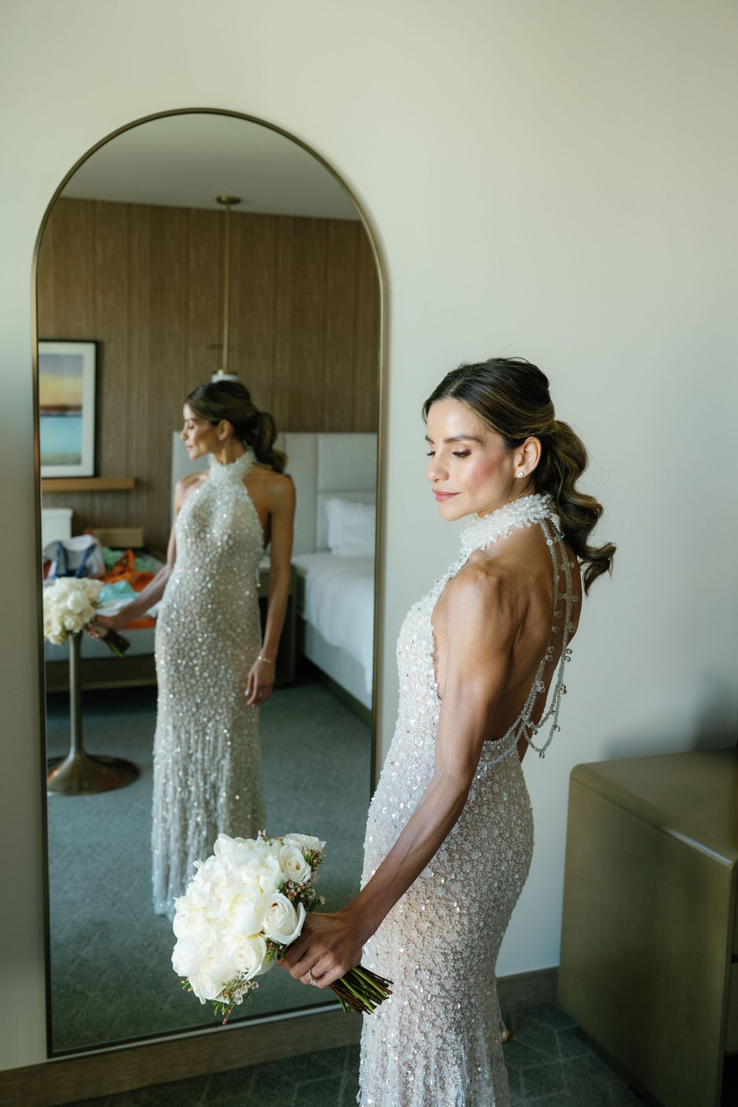 A bride in a beaded white gown stands next to a mirror holding a white rose bouquet in an elegant dressing room.