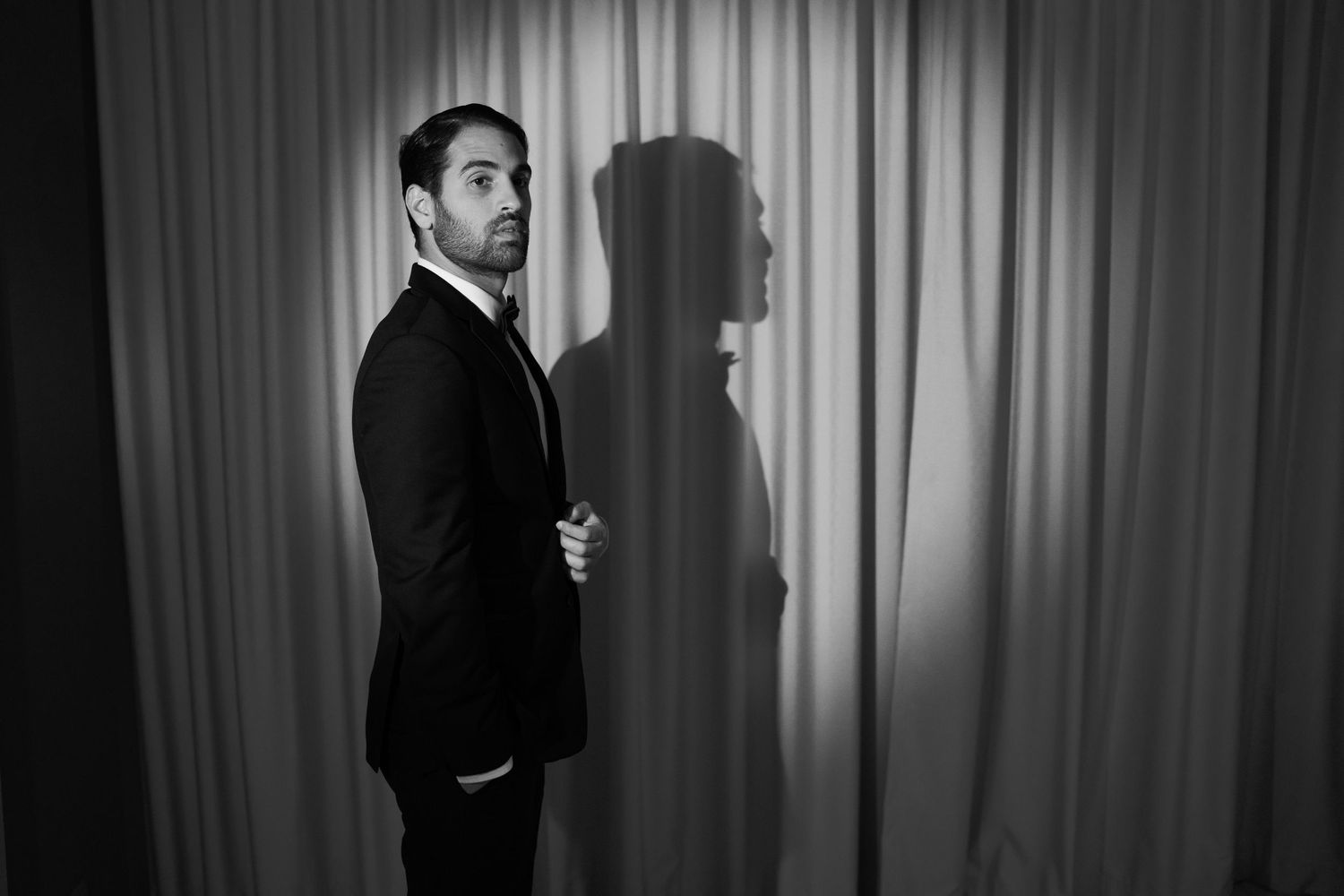 A dramatic black and white portrait of a groom in a suit casting a shadow against light curtains.