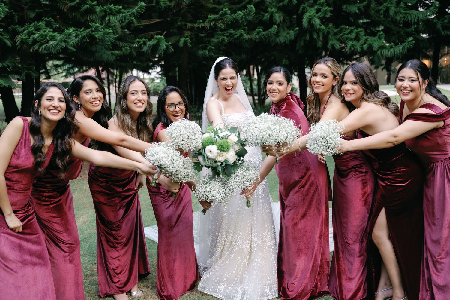 Wedding party in burgundy dresses and white gown pose together outdoors for group photos.