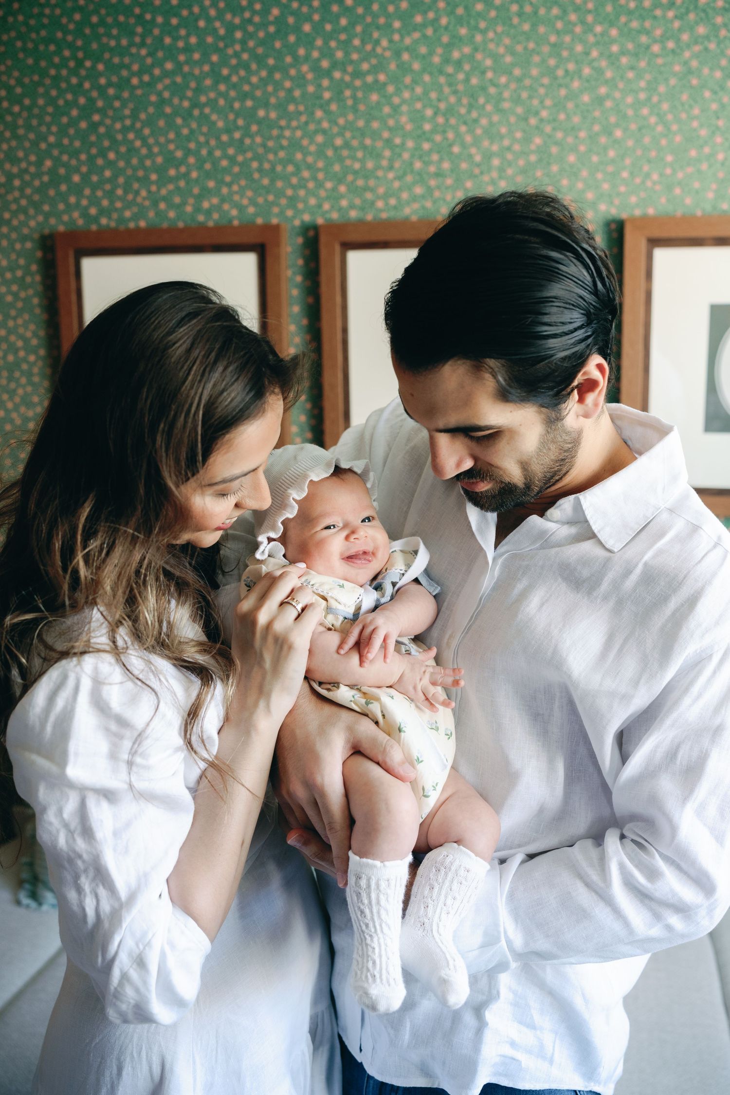 A couple in white shirts lovingly gazes at their newborn baby, held gently against a green patterned wall.