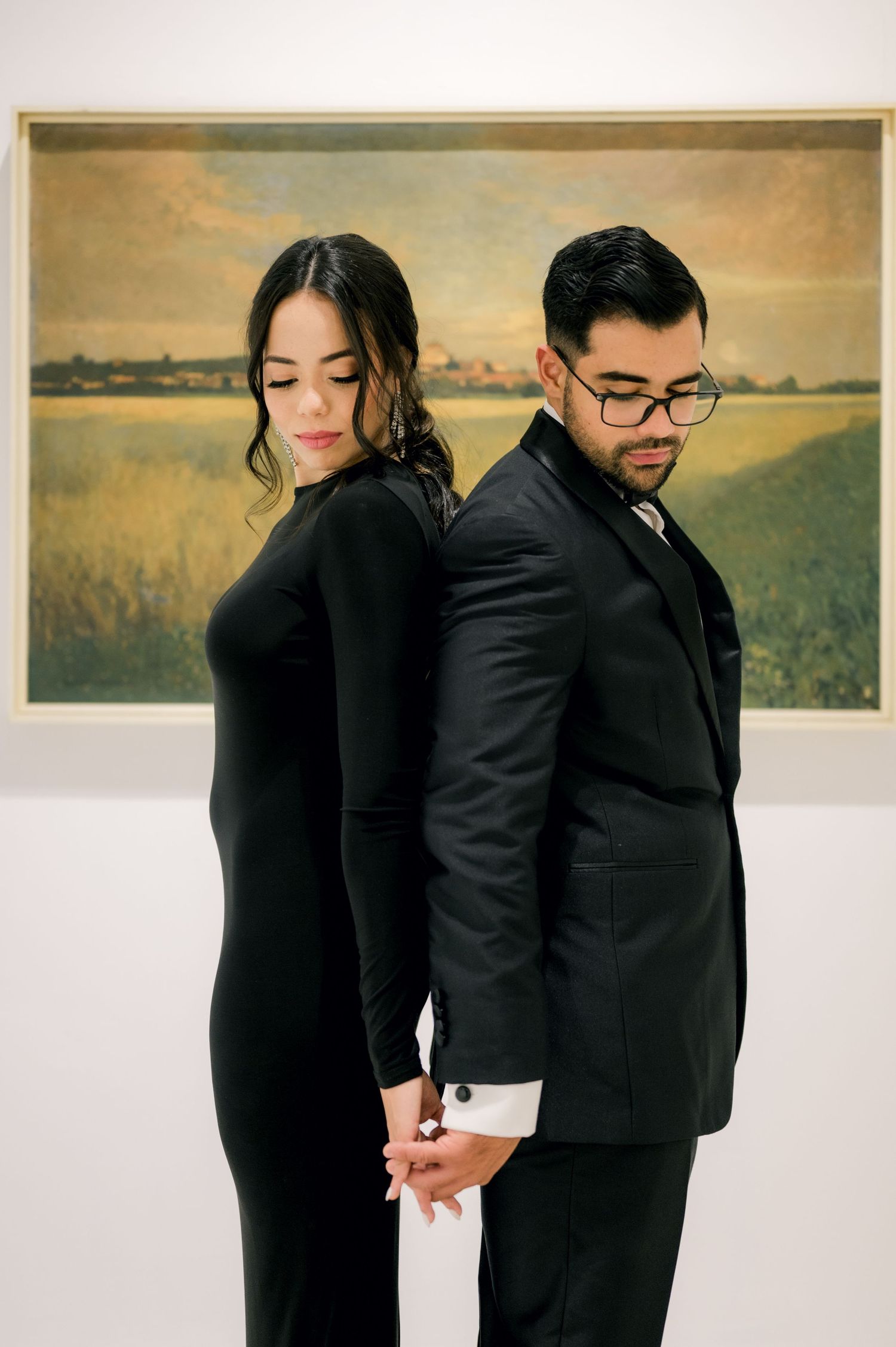 Elegant couple in black formal attire stand back-to-back in front of a landscape painting in an art gallery.